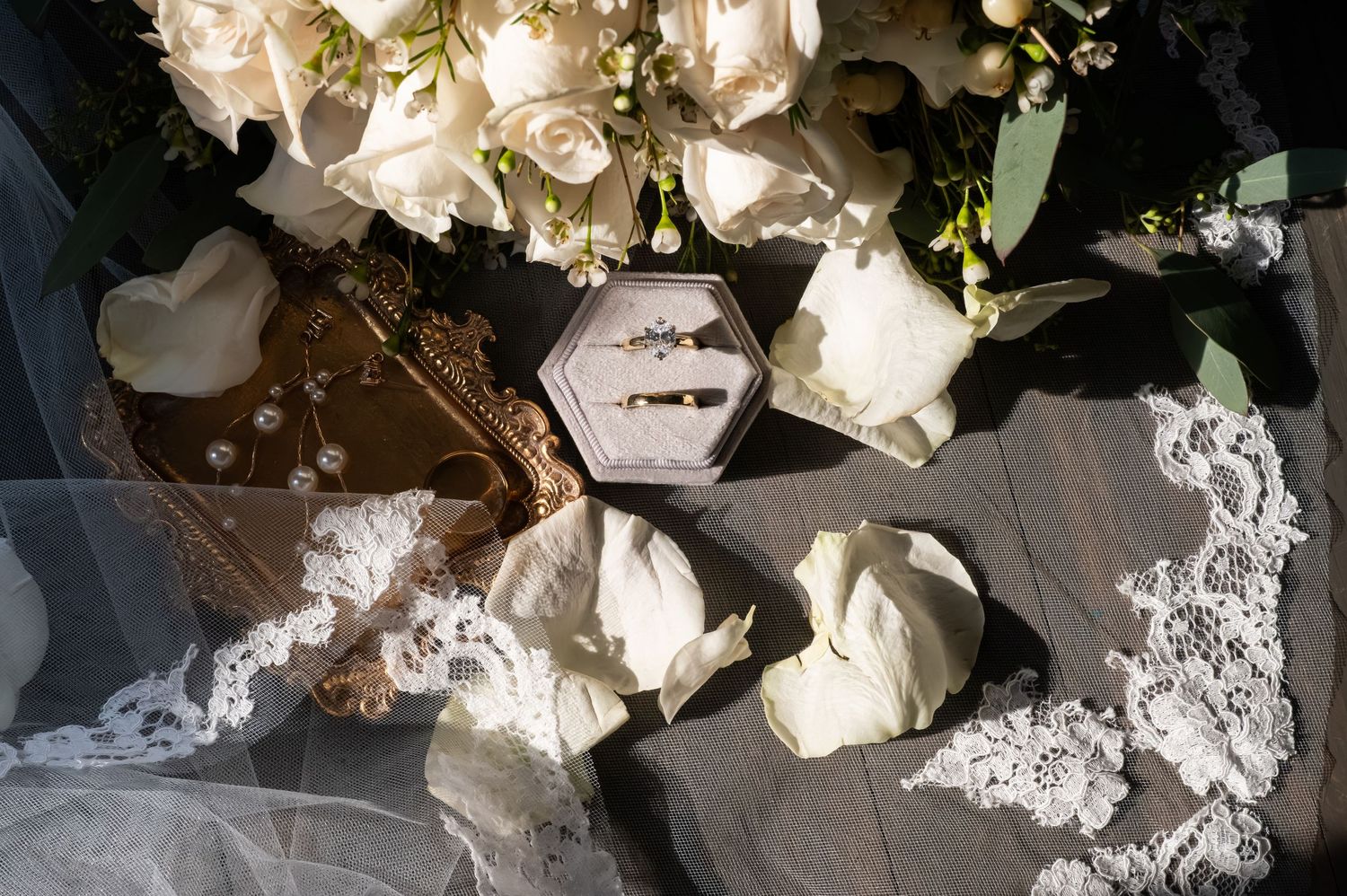 Elegant wedding flat lay with white roses, lace veil, gold ring box, and diamond rings on rustic wood surface.