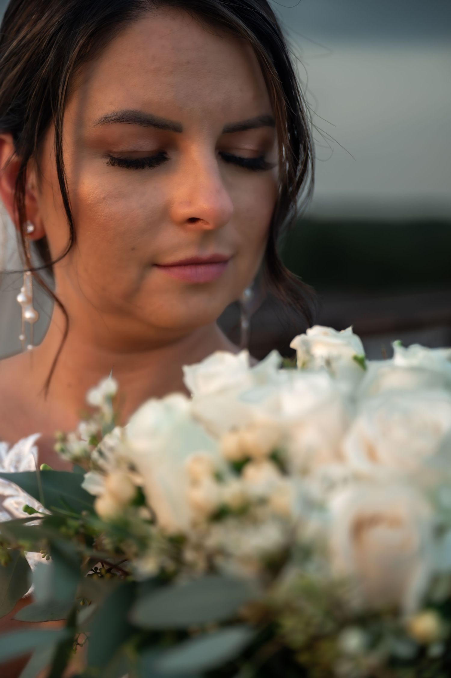 Bride gazing down at elegant white rose bouquet with greenery, wearing pearl earrings and soft bridal makeup.