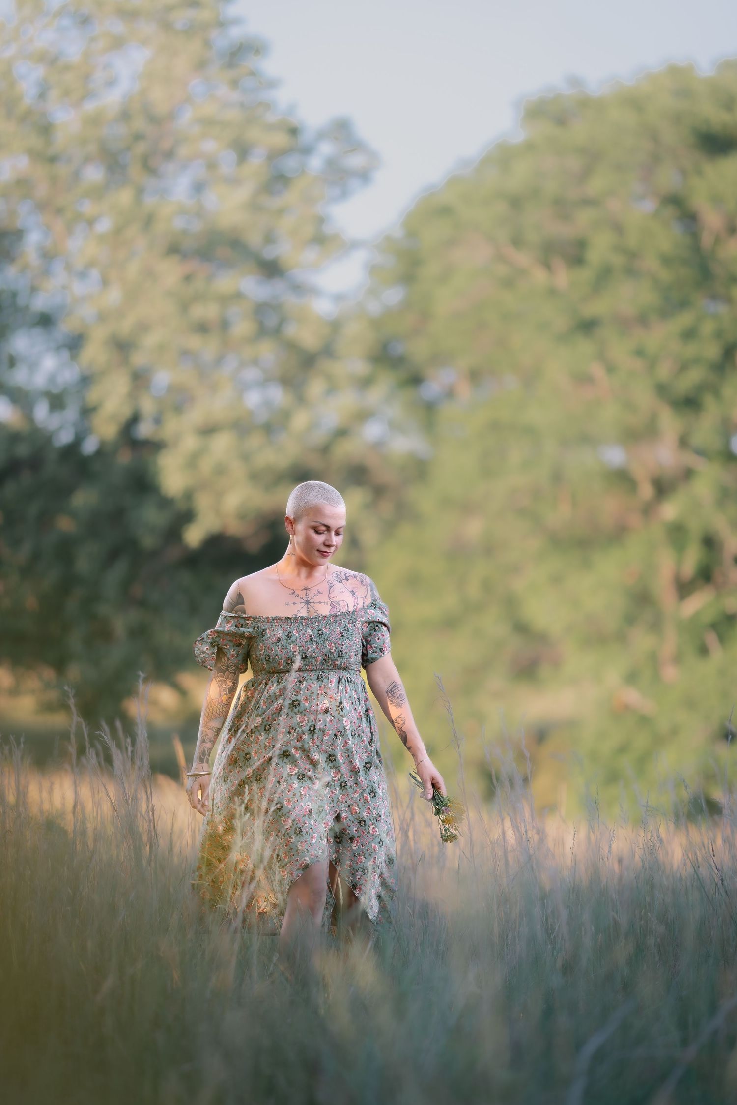 Woman in floral dress walking through tall golden grass field surrounded by green trees.