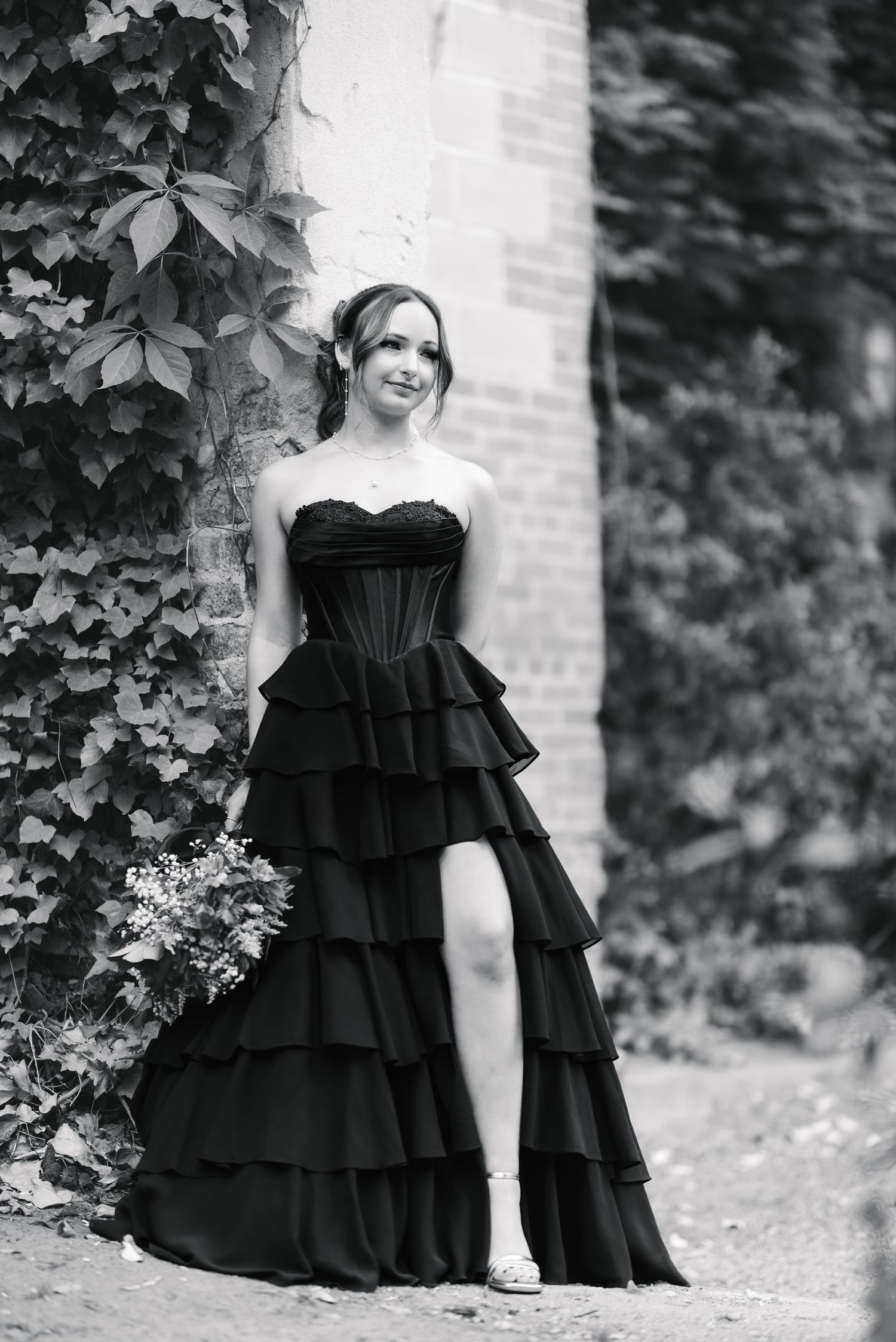 Young woman in elegant black strapless ruffled gown posing outdoors near ivy-covered stone wall in black and white photo.