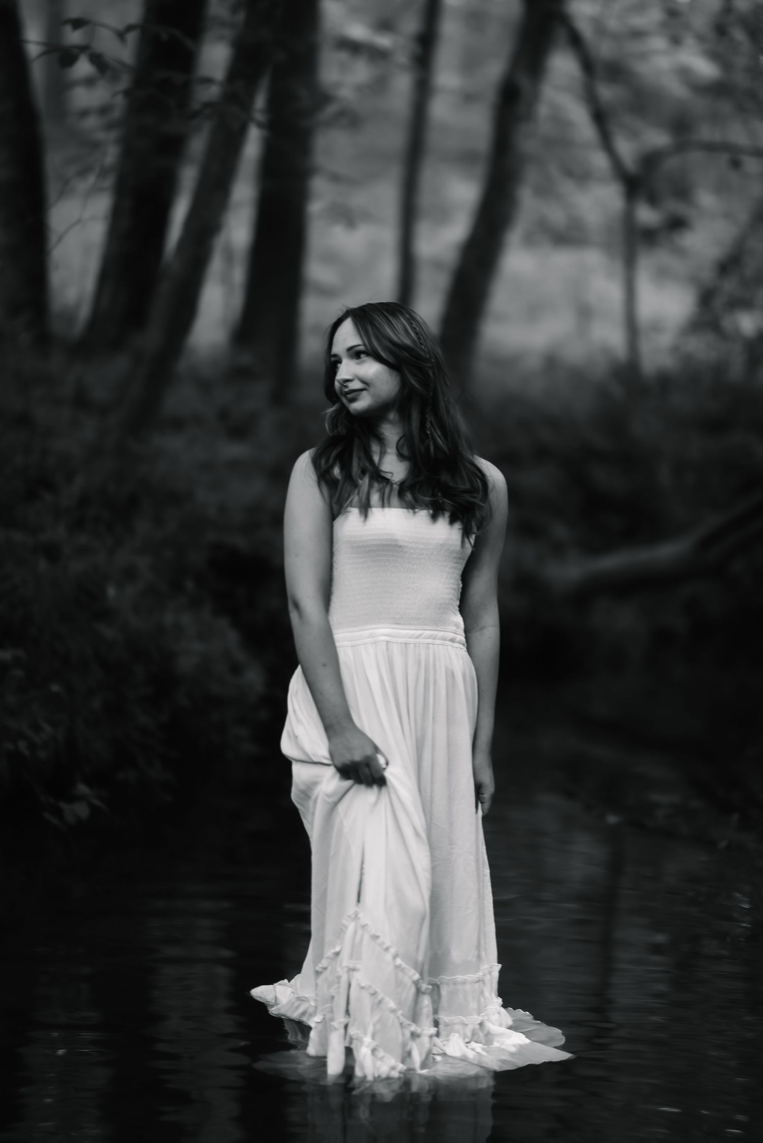 Black and white photo of a woman in a white dress standing in a forest stream, smiling softly.