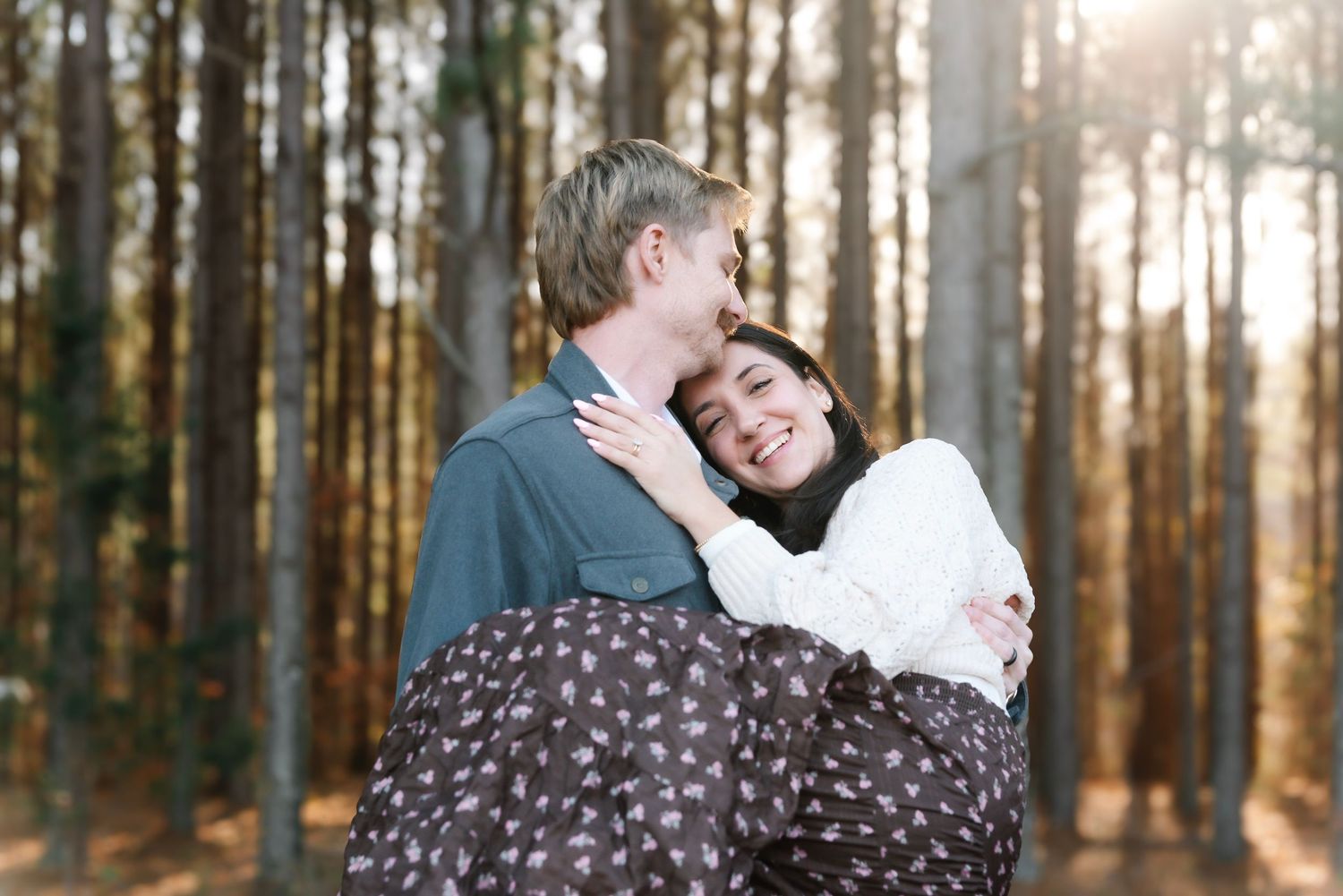 Couple embracing romantically in a sunlit pine forest, woman smiling warmly while being held by her partner.