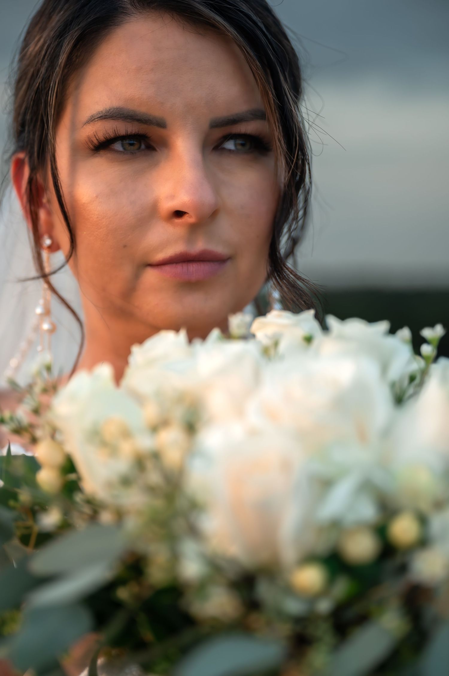 Bride holding white rose bouquet with elegant updo hairstyle and pearl earrings on her wedding day.
