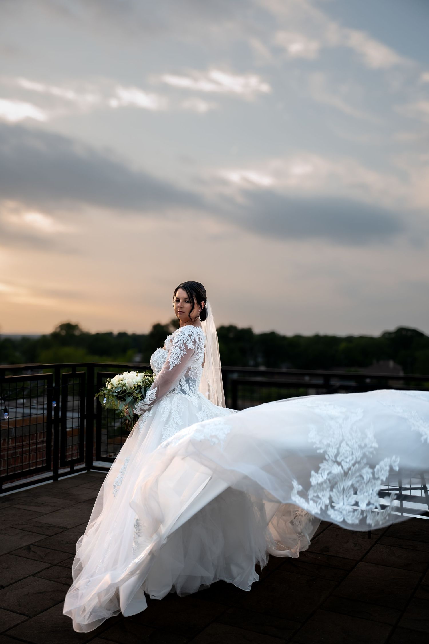 Bride in flowing white lace gown with veil billowing in wind holds bouquet on rooftop terrace at sunset.