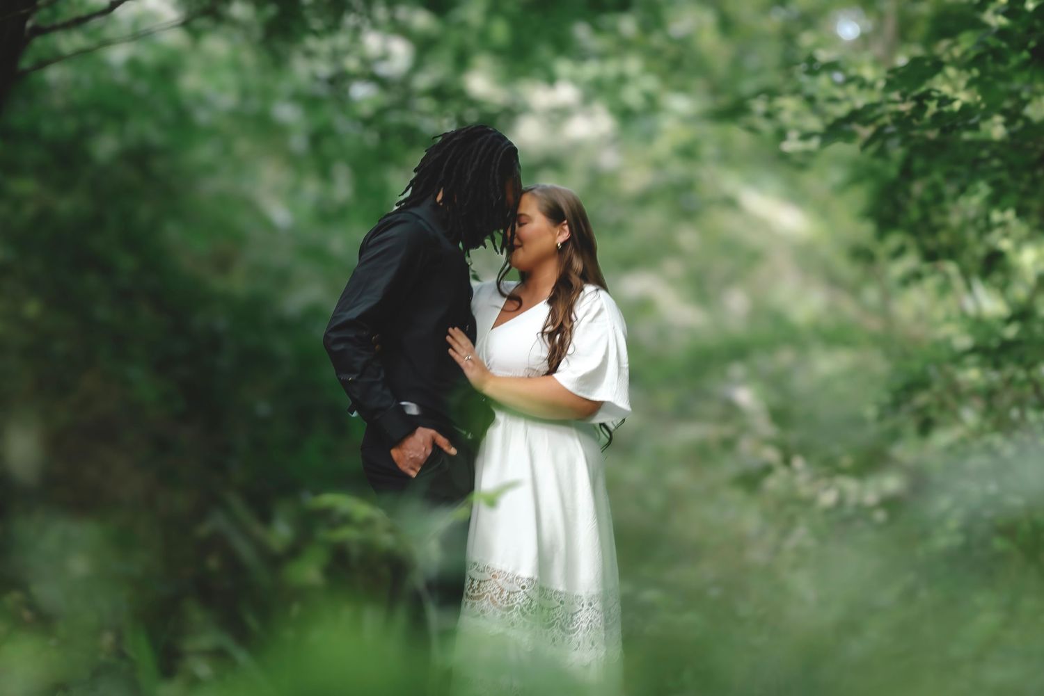Couple sharing a romantic moment surrounded by lush green foliage, woman in white dress and man in black suit.