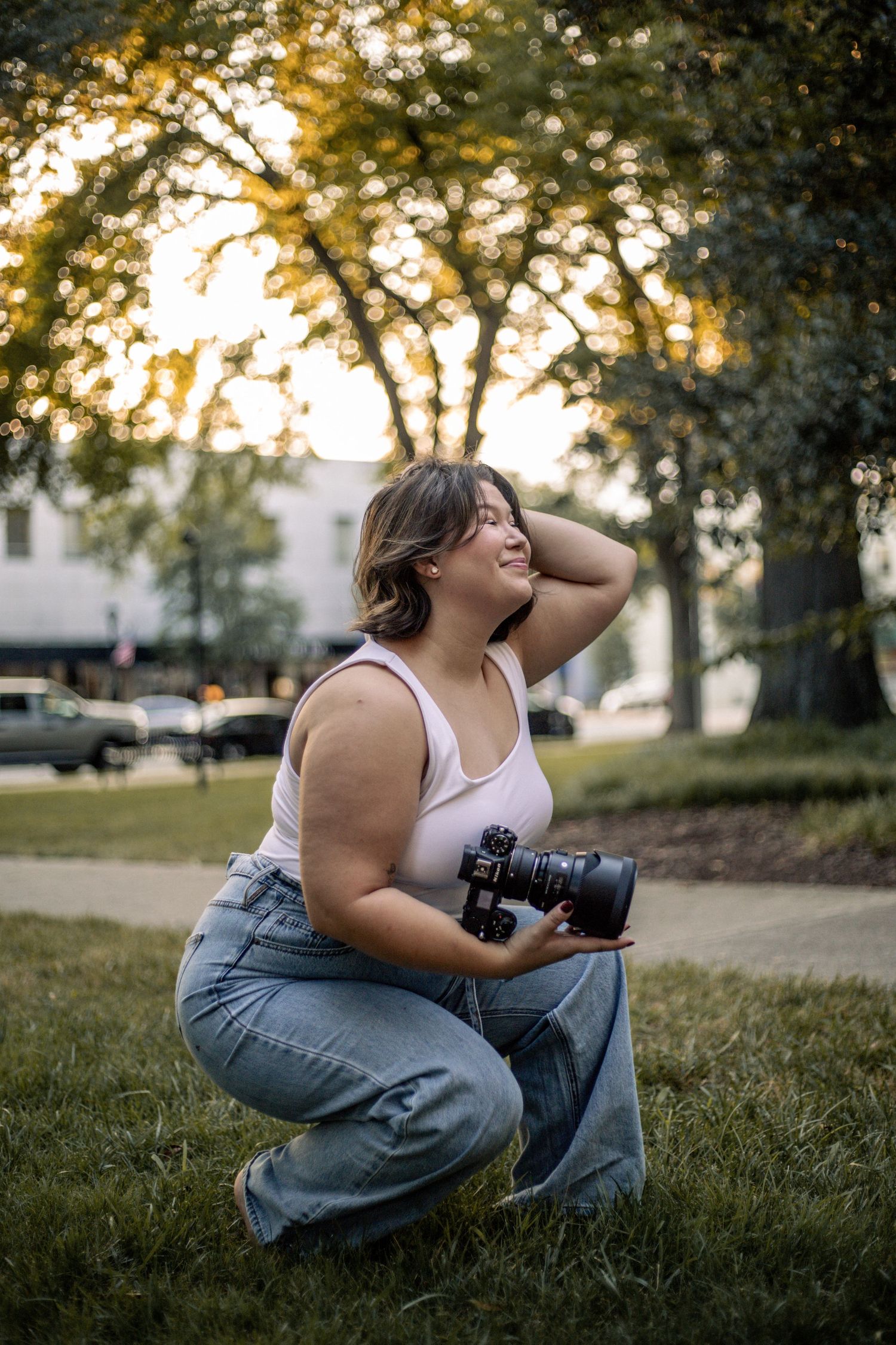 Woman crouching on grass holding a camera, smiling outdoors with trees and warm golden hour light in the background.