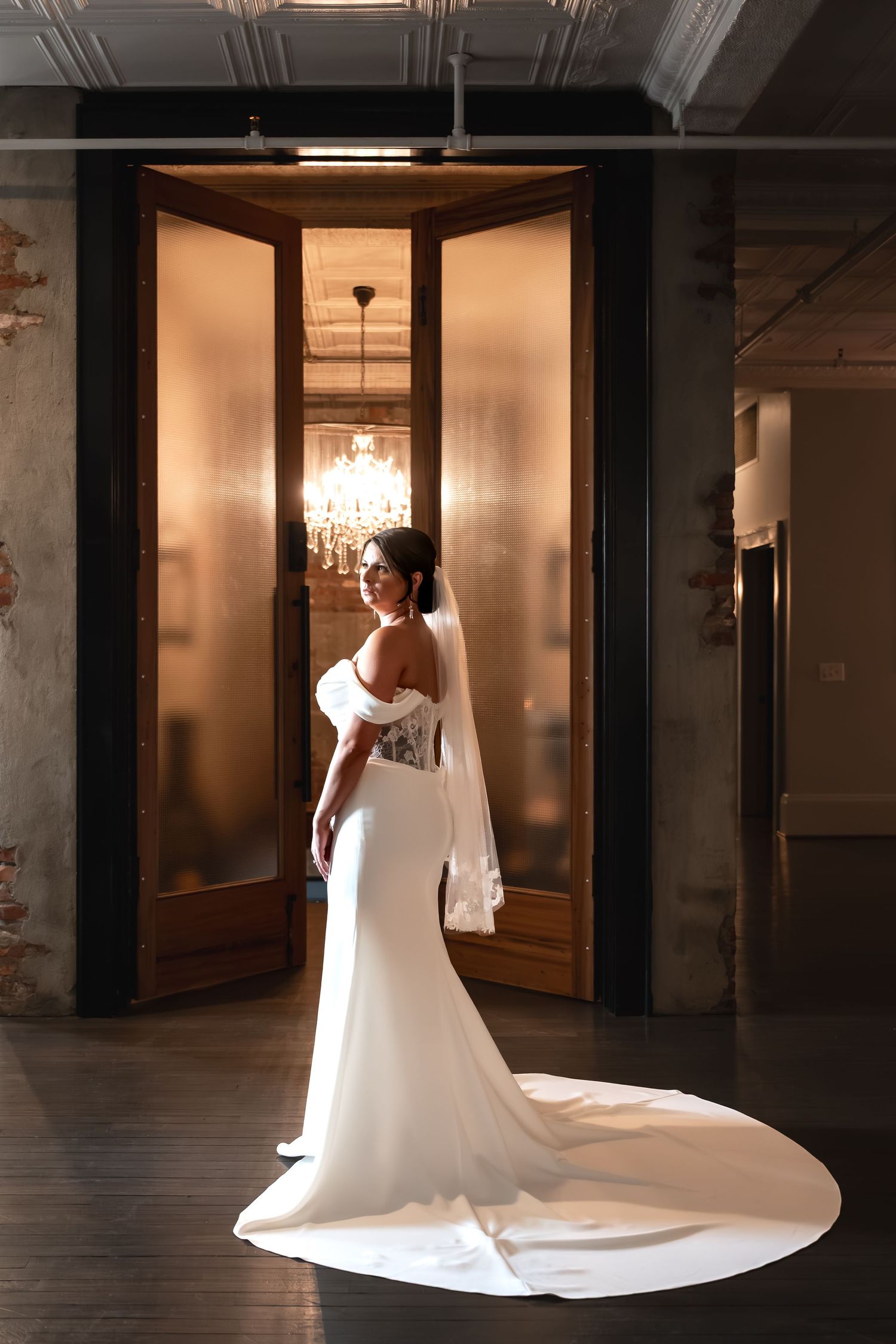 Bride in elegant white off-shoulder gown with long train stands before ornate wooden doors with chandelier backdrop.