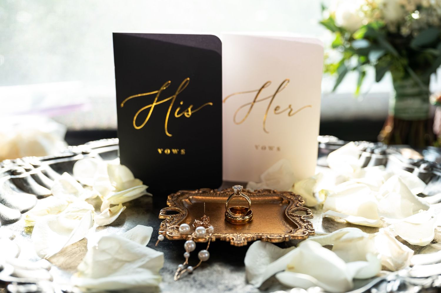 His and Her gold foil vow books with wedding rings on ornate tray surrounded by white rose petals.