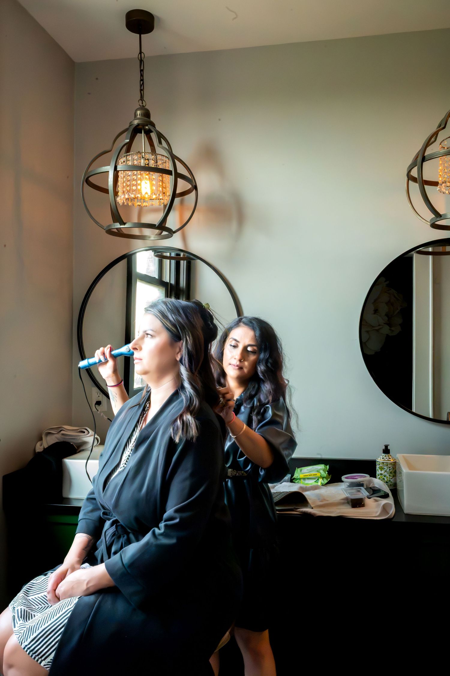 A makeup artist applies makeup to a woman in a black robe, seated in a stylish salon with round mirrors and pendant lights.