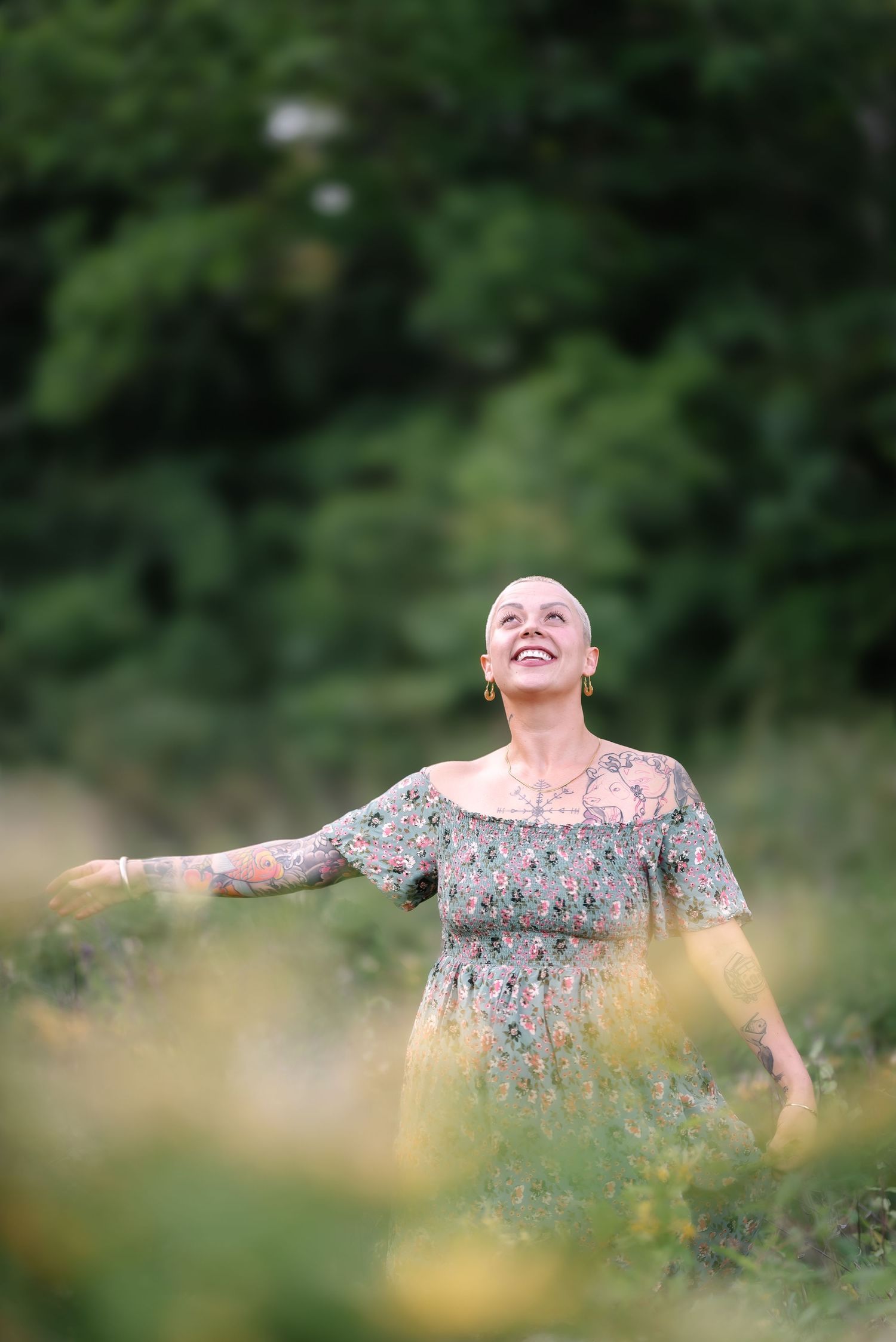Bald woman in floral dress smiling joyfully outdoors in a lush green field, arms outstretched in celebration.