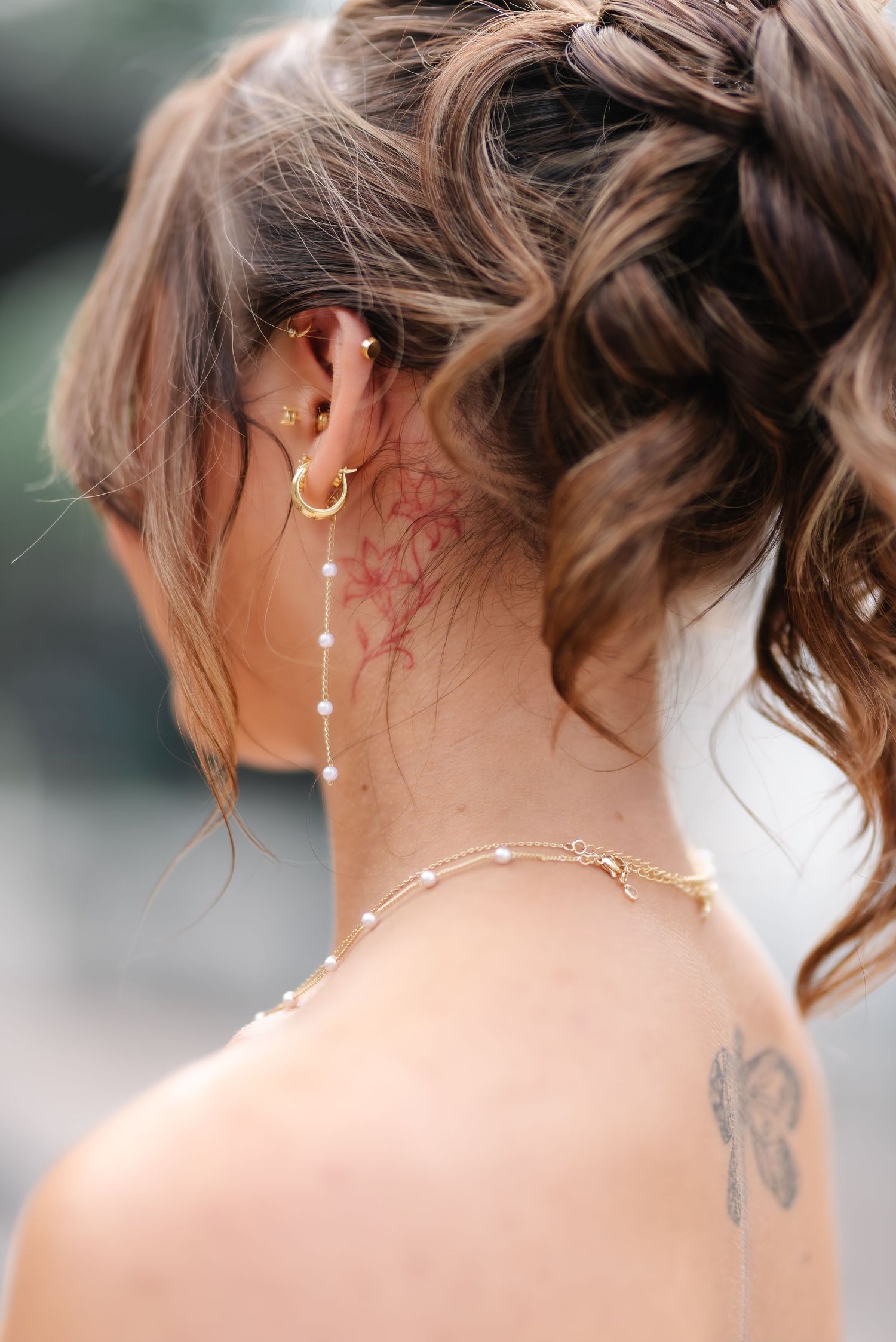 Woman with braided updo, gold hoop earrings, pearl chain, and floral neck tattoo, viewed from behind.