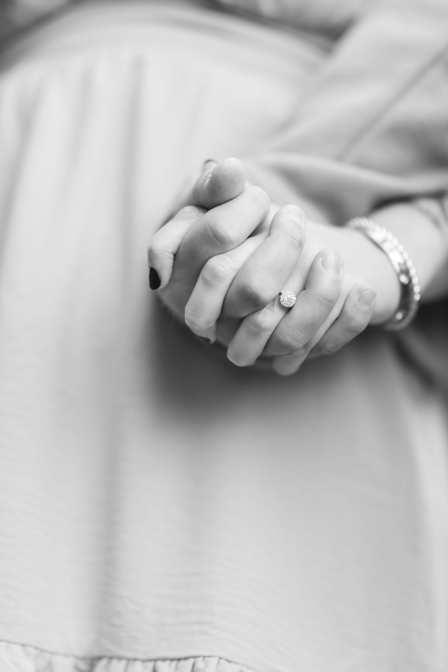 Black and white close-up of clasped hands wearing diamond ring and bracelet over soft fabric.