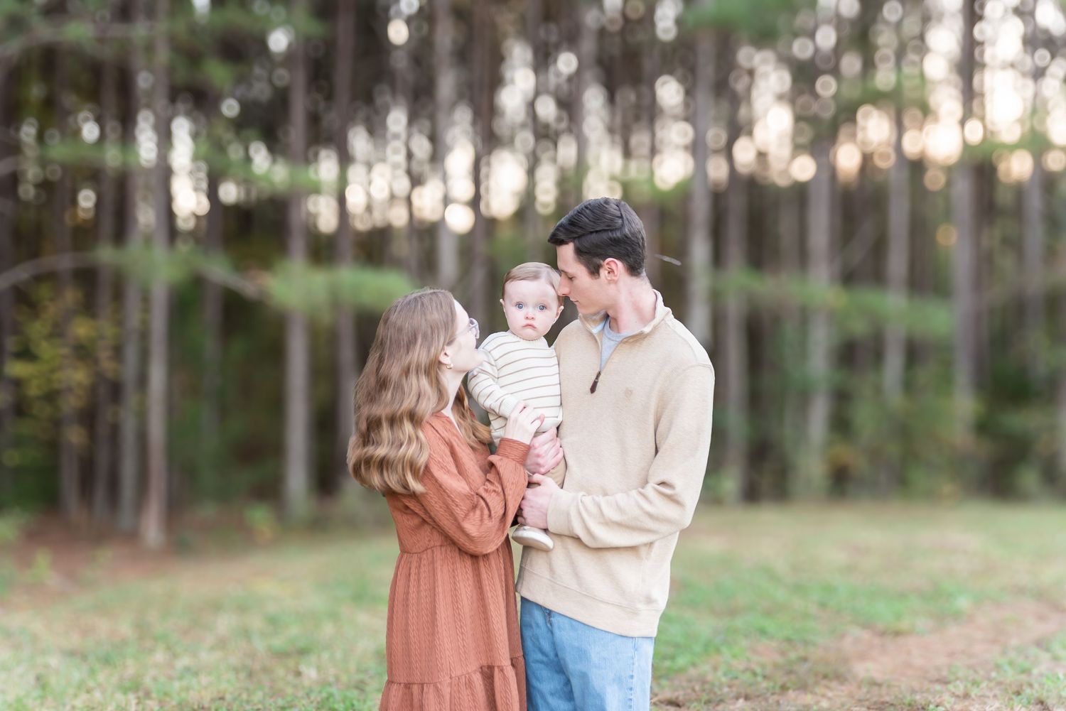 A young family with a baby poses in a sunlit forest clearing, mom in rust dress, dad in beige sweater and jeans.