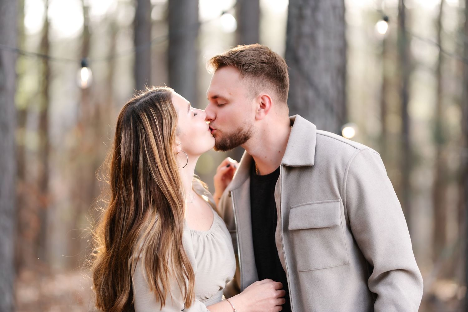 Couple sharing a romantic kiss in a pine forest, wearing casual outfits in a soft bokeh outdoor setting.