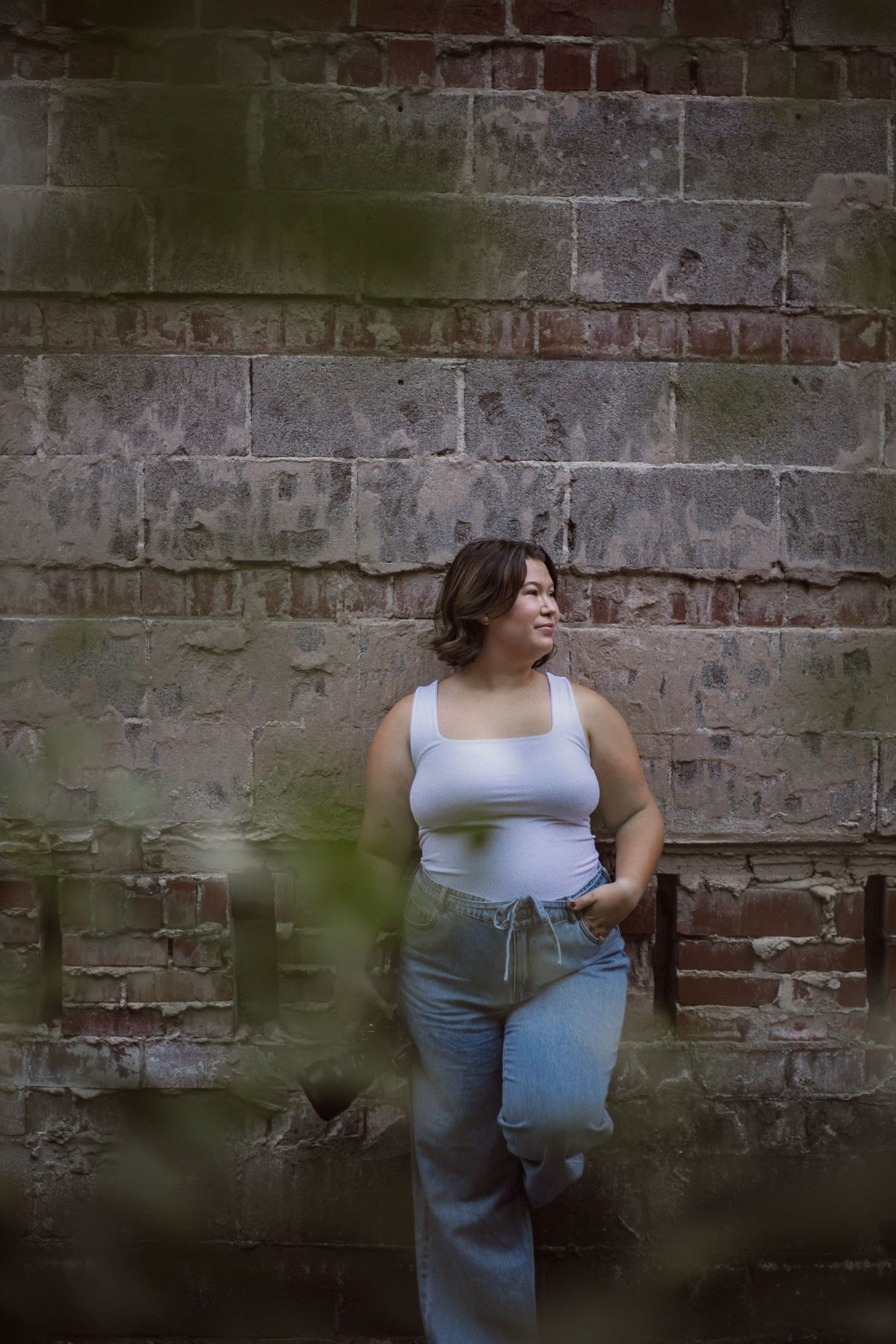 Young woman in white tank top and jeans leaning against a rustic brick wall, smiling and looking to the side.