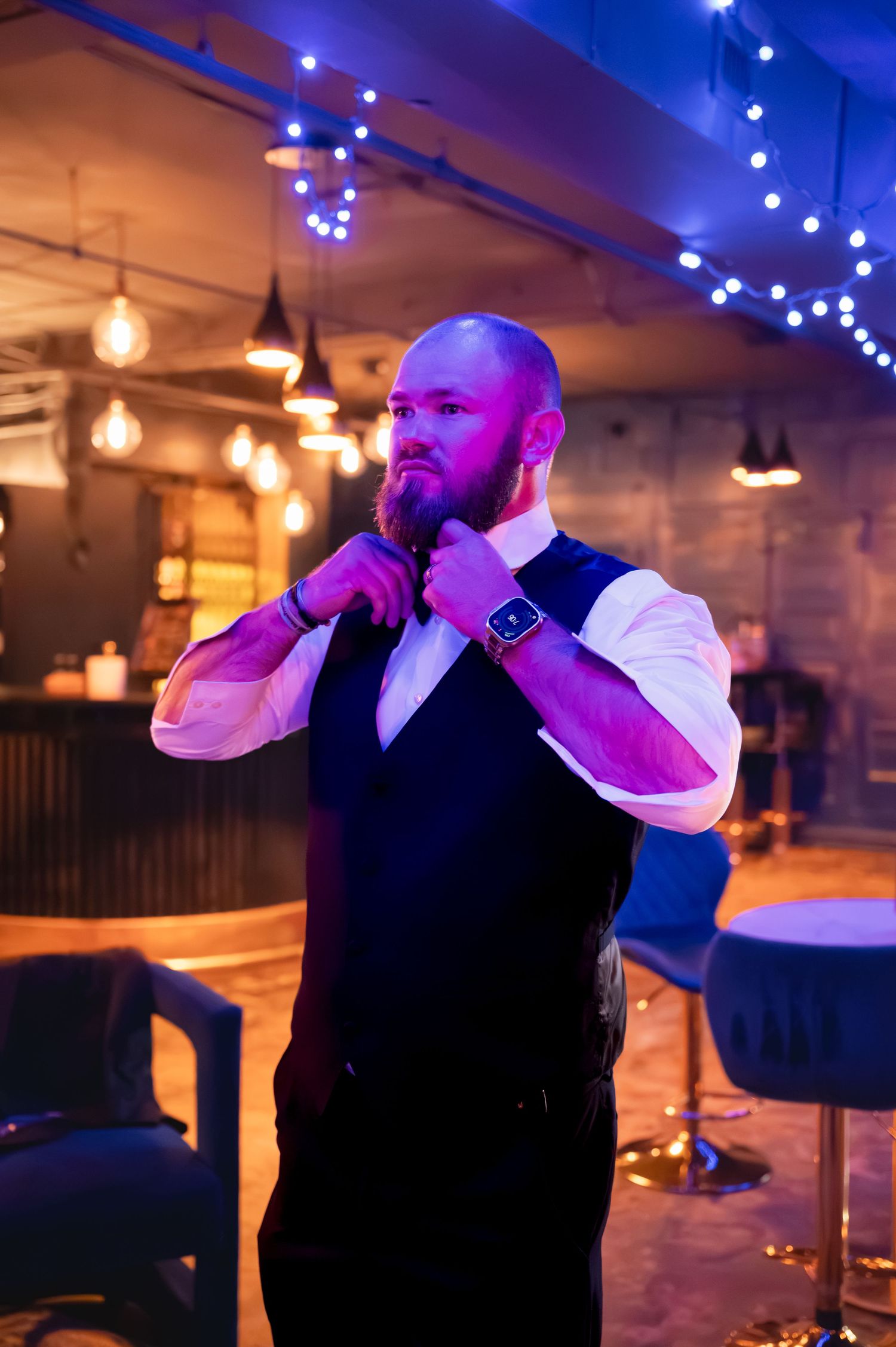 Bald bearded man in vest and white shirt adjusting tie in a bar with blue ambient lighting.