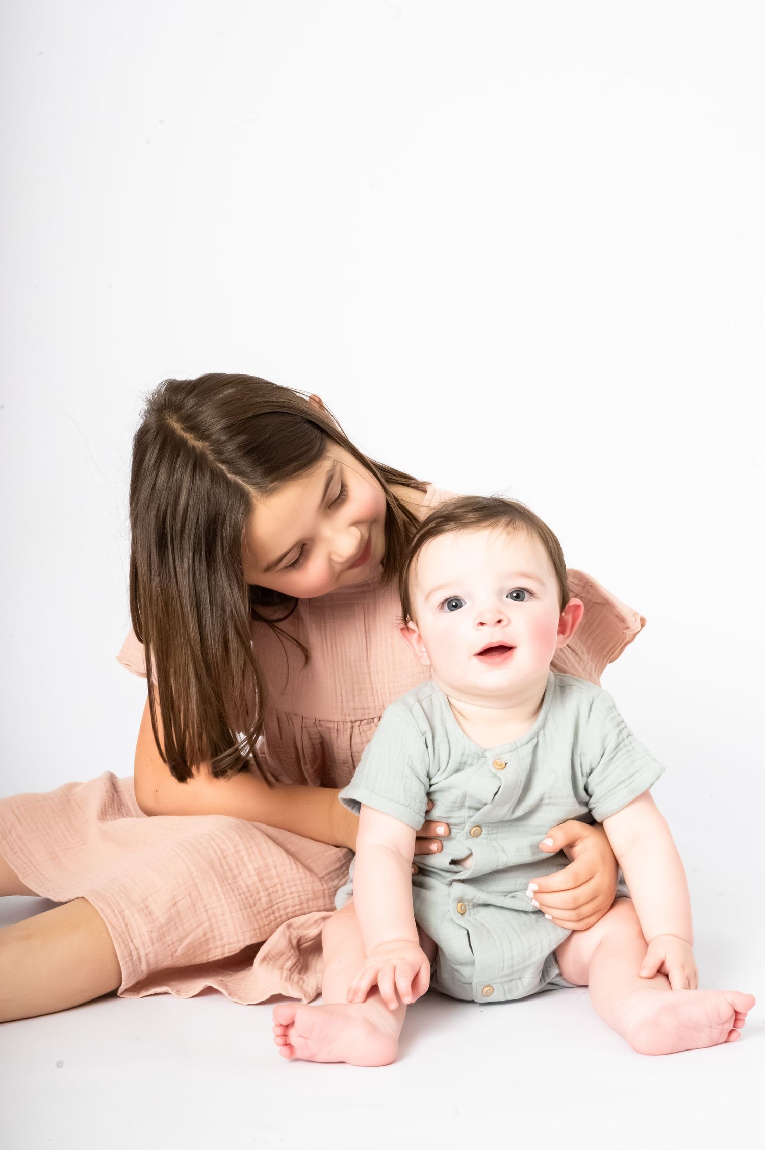 A young girl in a pink dress sits with a smiling baby in a gray romper against a white background.