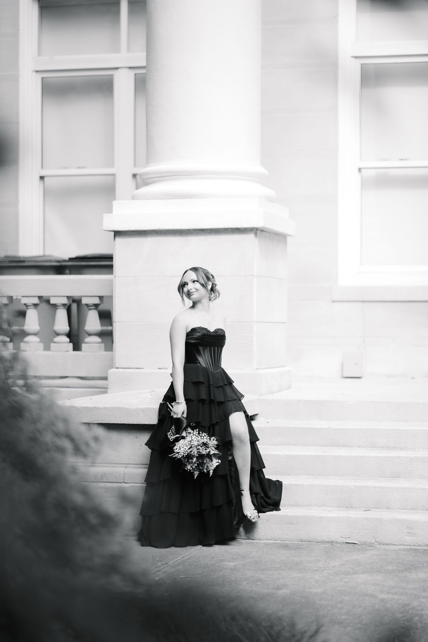 Woman in black strapless tiered gown holding bouquet poses on grand building steps in elegant black and white photo.