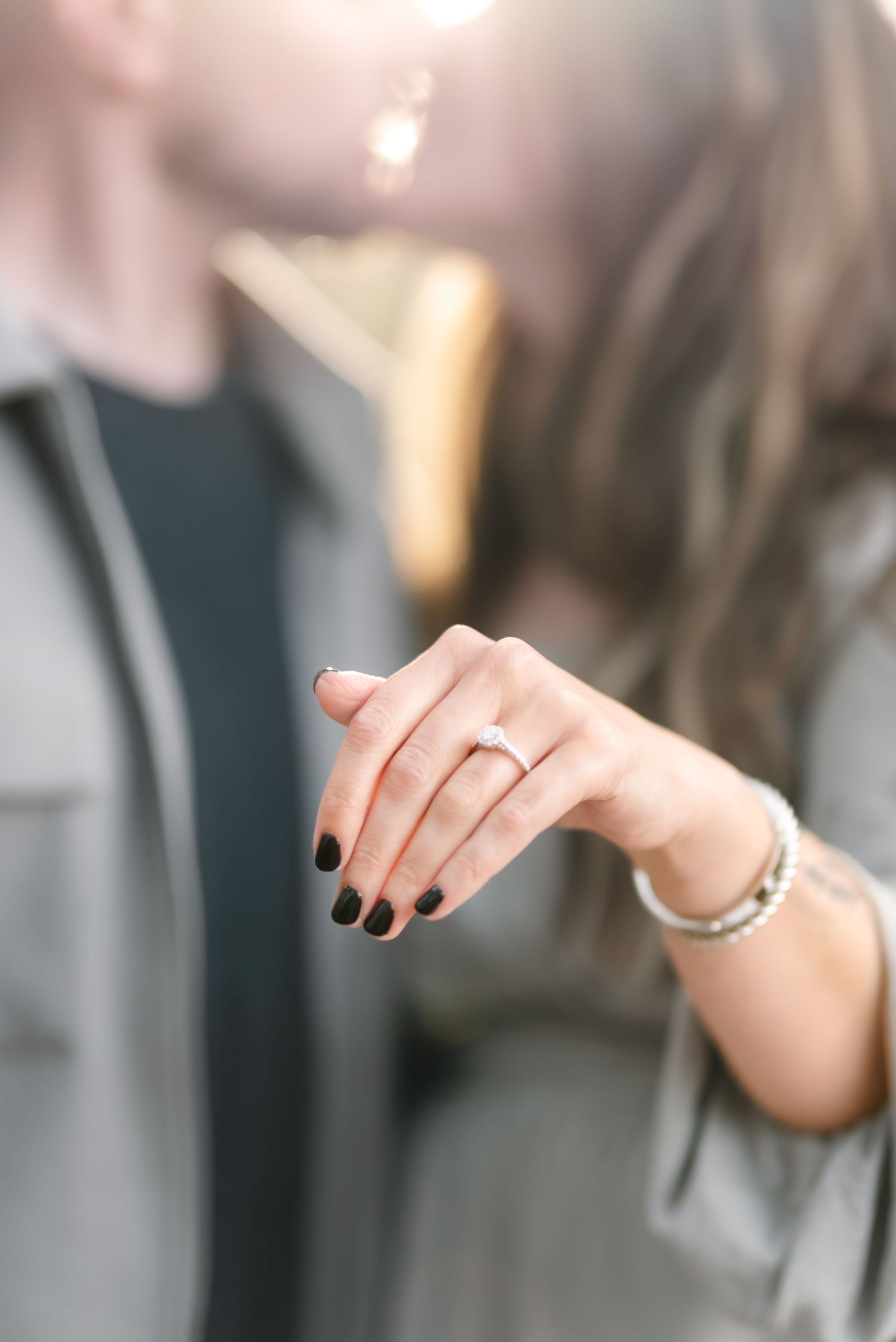 Woman showing off engagement ring with diamond solitaire, black nails, and bracelet while couple kisses in background.