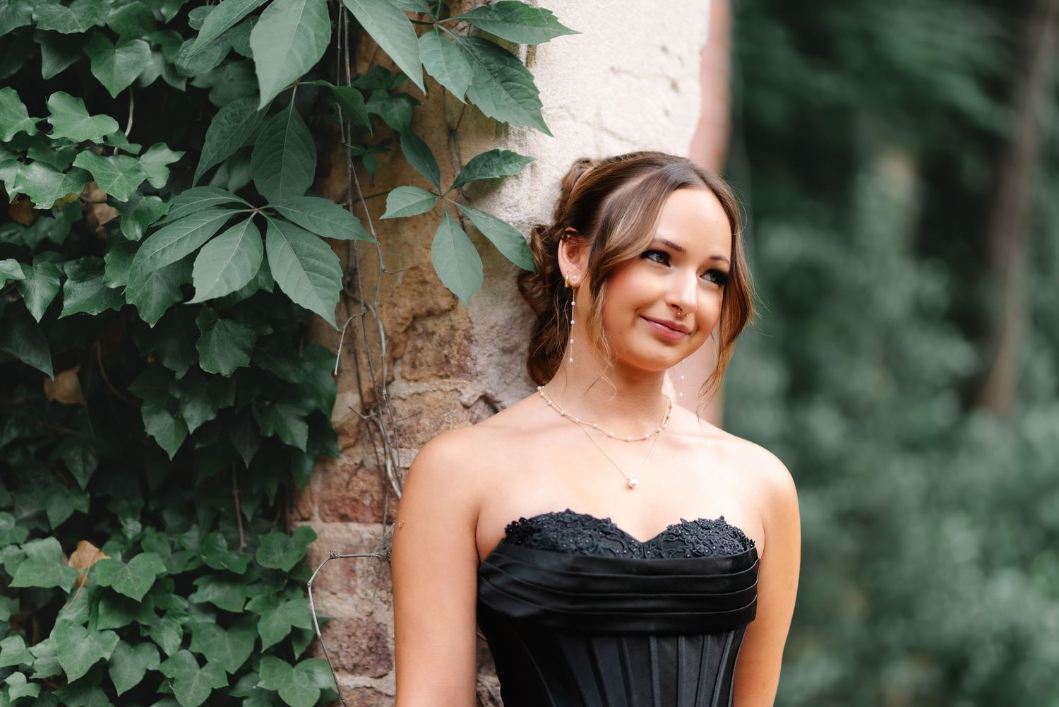 A woman in a black strapless gown smiles gracefully outdoors, posed against a stone wall with lush green ivy.