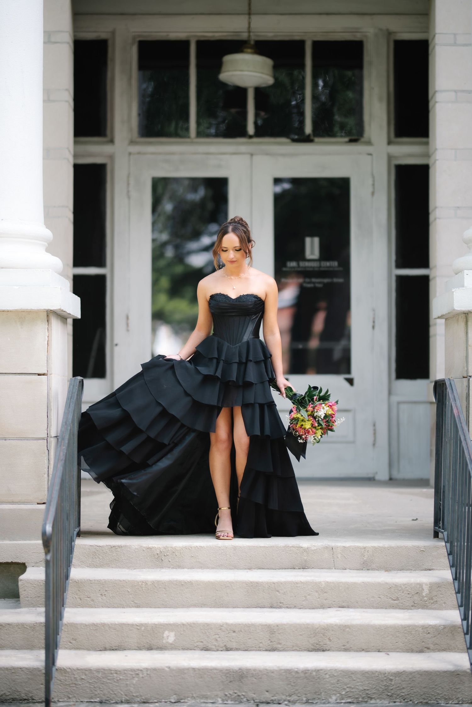 Woman in strapless black tiered high-low gown holding colorful bouquet descending stone steps at elegant venue entrance.