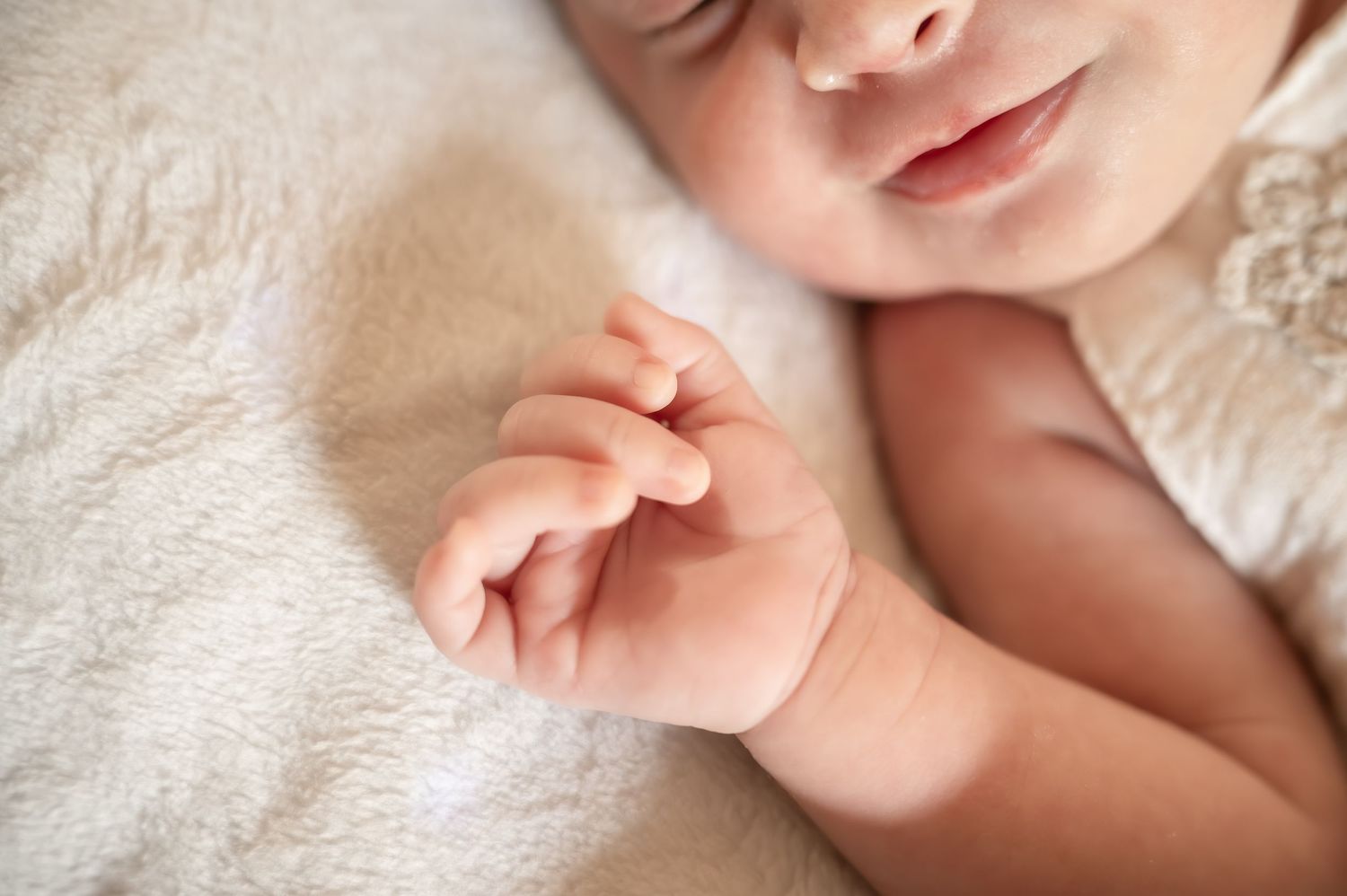 Close-up of a sleeping newborn baby's tiny hand and peaceful smile resting on a soft white blanket.