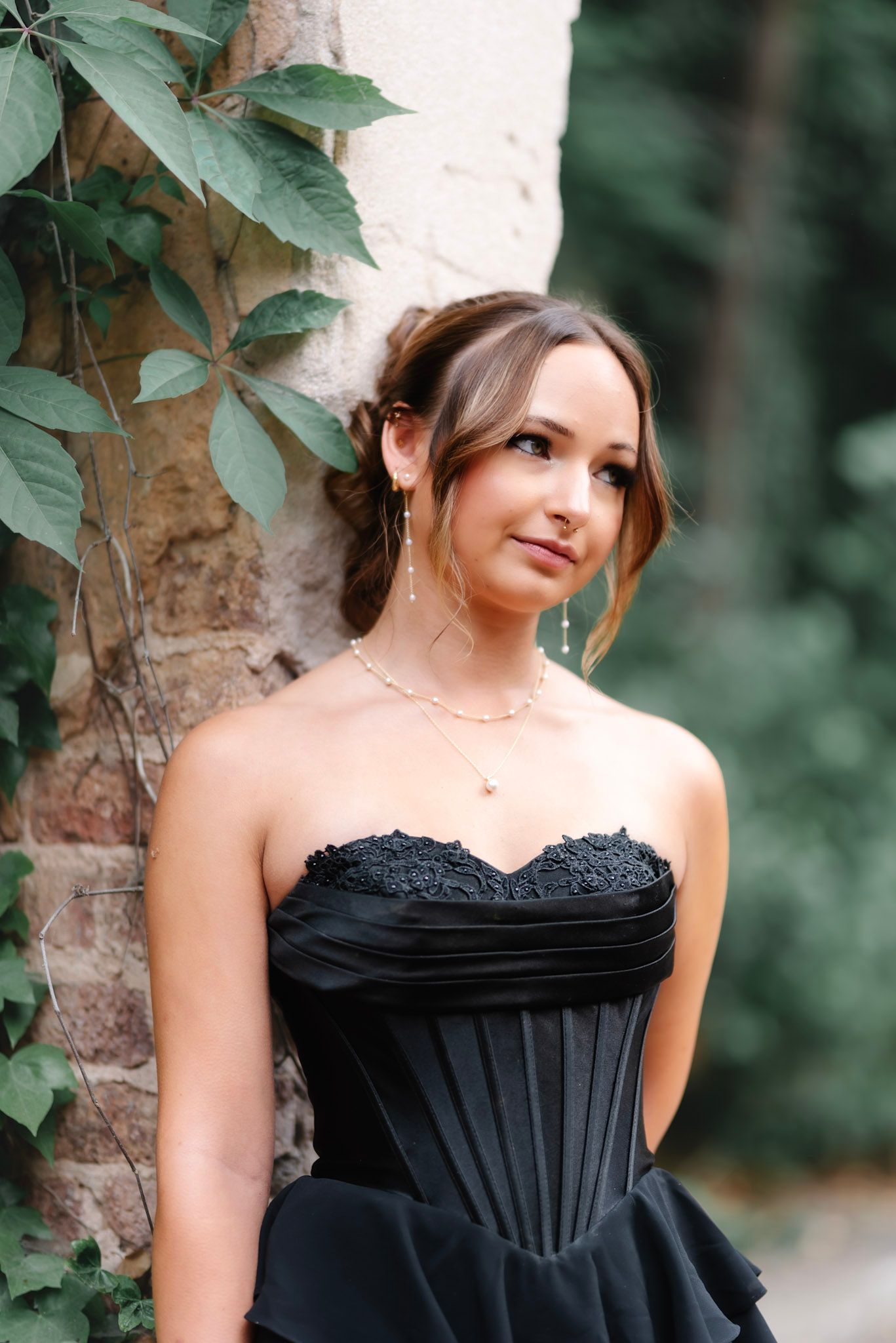 Young woman in elegant black strapless corset gown with lace detail, gold necklace, updo hairstyle, posing outdoors.