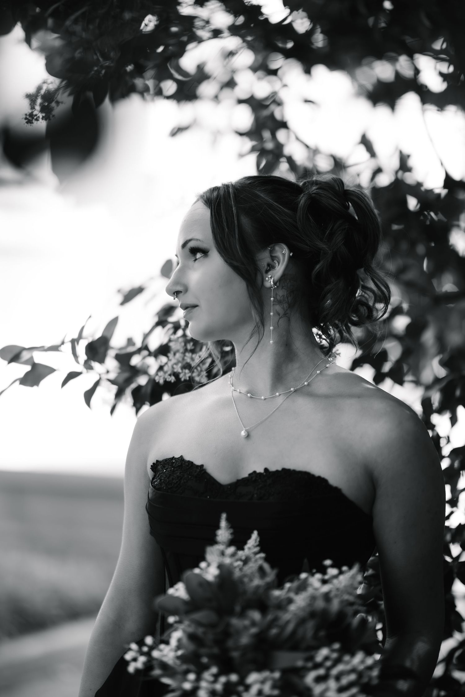 Black and white portrait of a bride in strapless gown with updo hairstyle and pearl necklace holding bouquet outdoors.