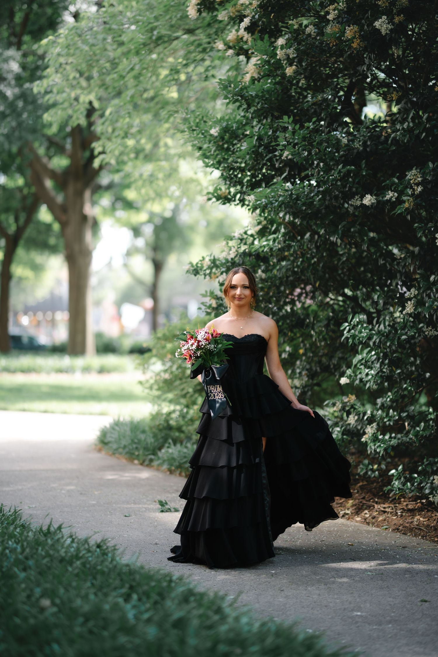 Bride in a strapless black tiered ballgown holding a floral bouquet while walking along a tree-lined garden path.
