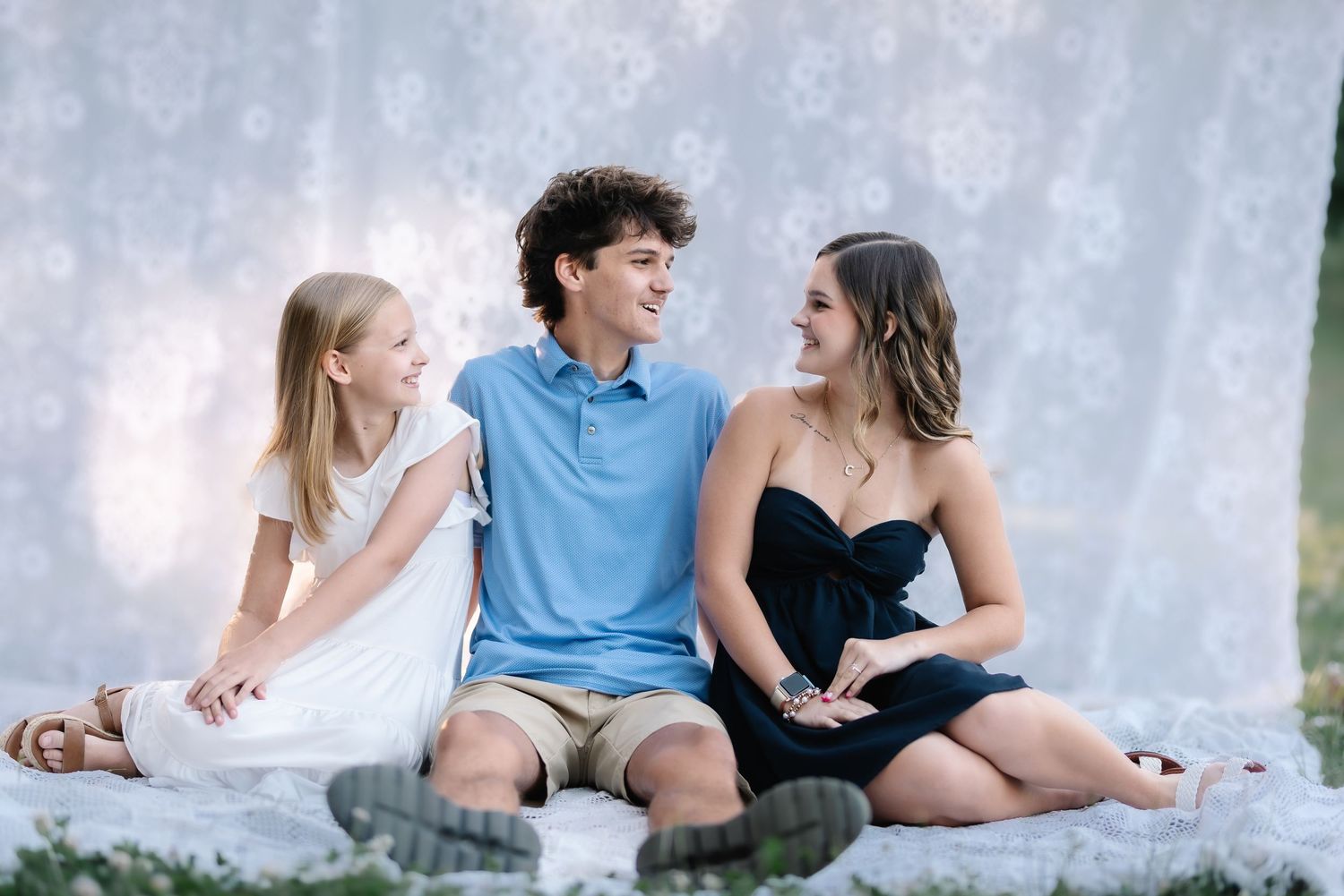 Three smiling teens sit together on a blanket outdoors in front of a floral backdrop for a portrait photo.
