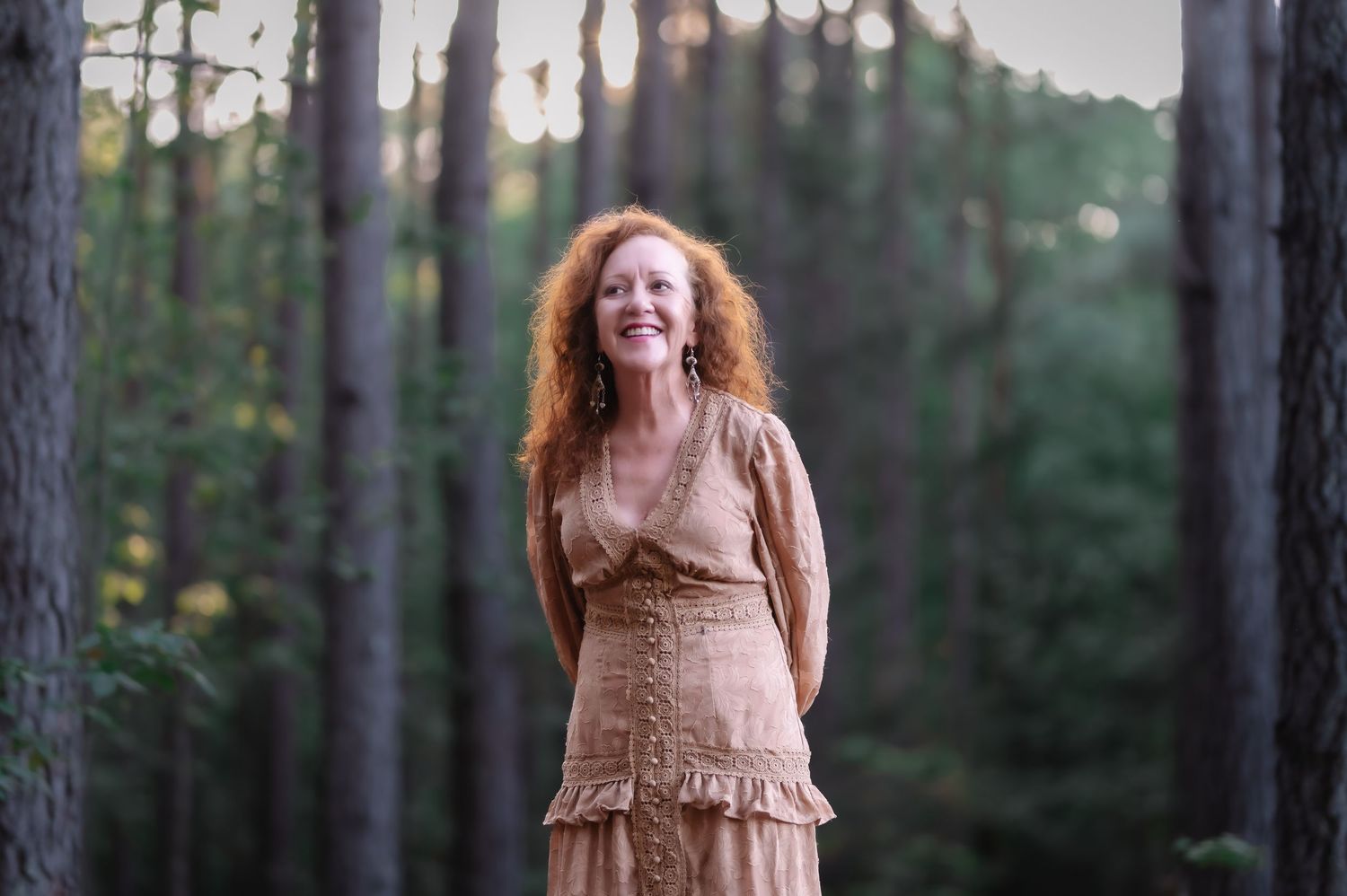 A smiling redhead woman in a beige boho dress stands joyfully among tall pine trees in a forest.