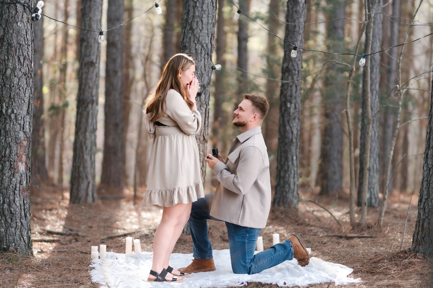 Man proposes to woman on one knee in a romantic forest setting with candles and string lights.