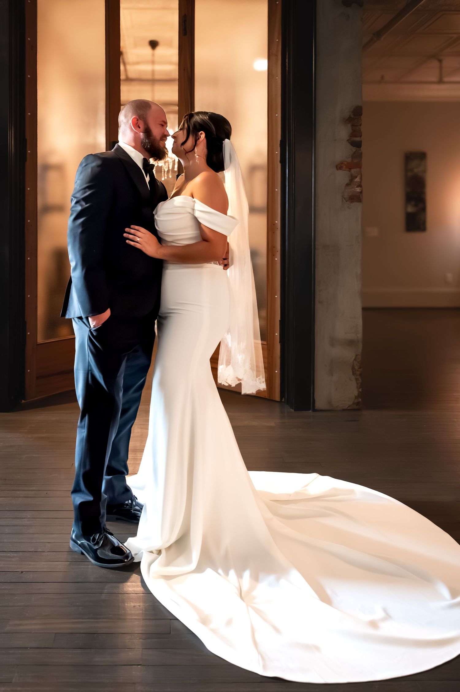 A bride and groom share a romantic kiss in an elegant indoor venue, her white off-shoulder gown with long train pooling gracefully.