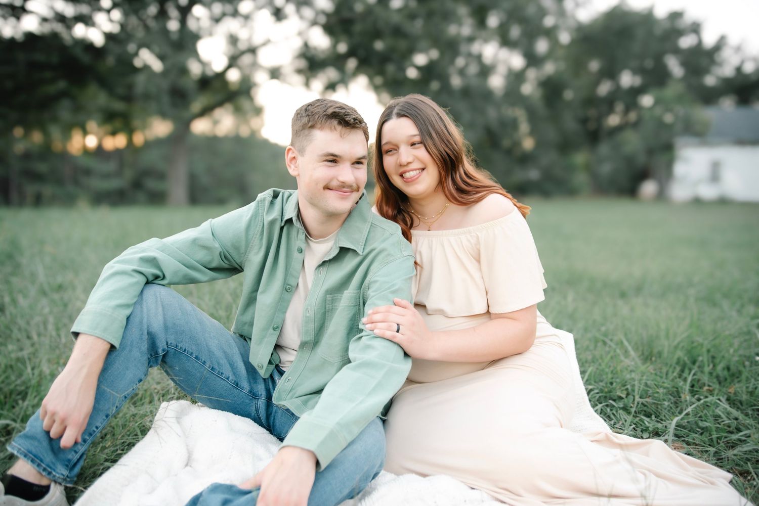 A smiling couple sits on a white blanket in a green field during golden hour, dressed in casual pastel outfits.