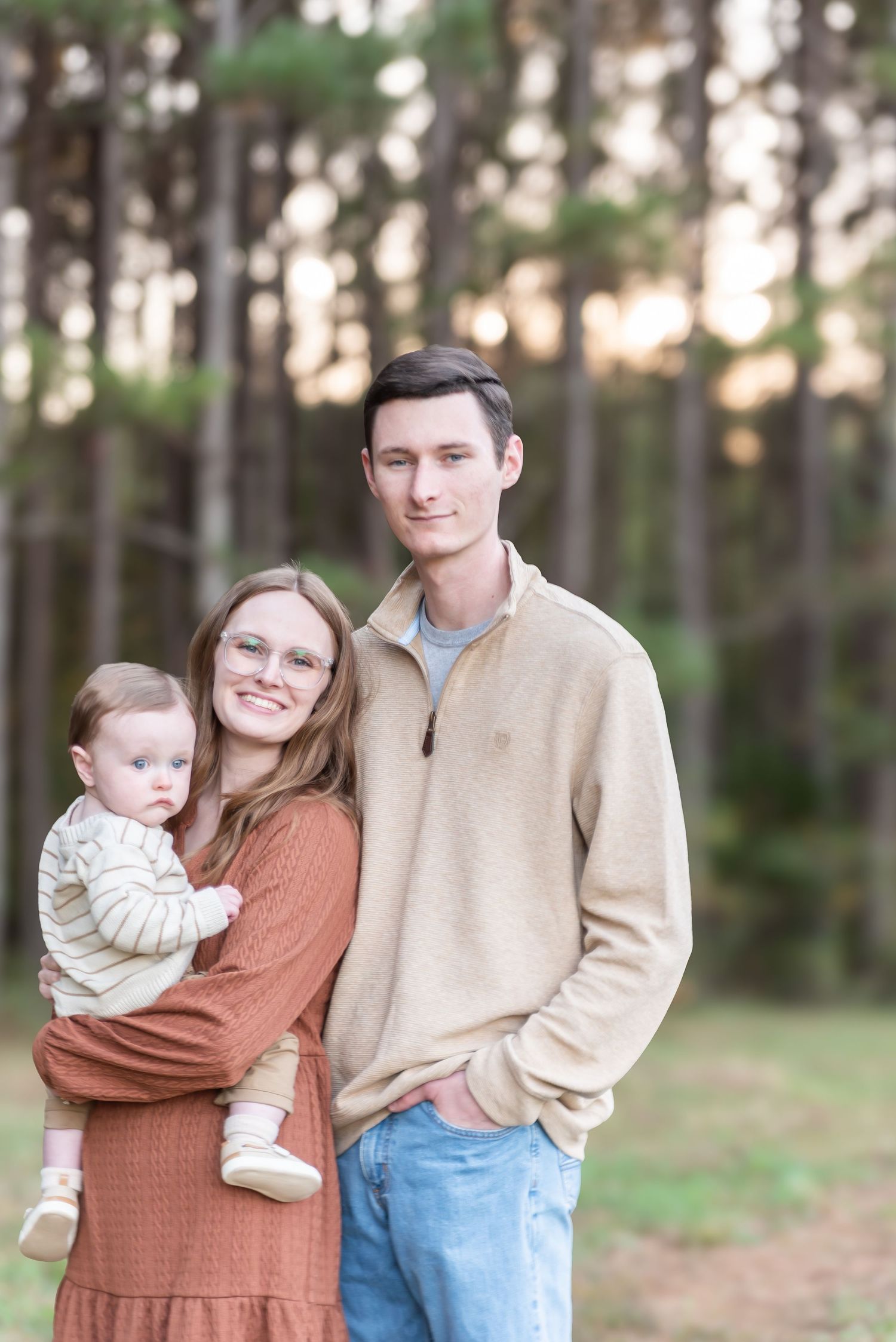 Young couple smiling outdoors in a pine forest, holding their baby dressed in a striped outfit, wearing casual fall attire.