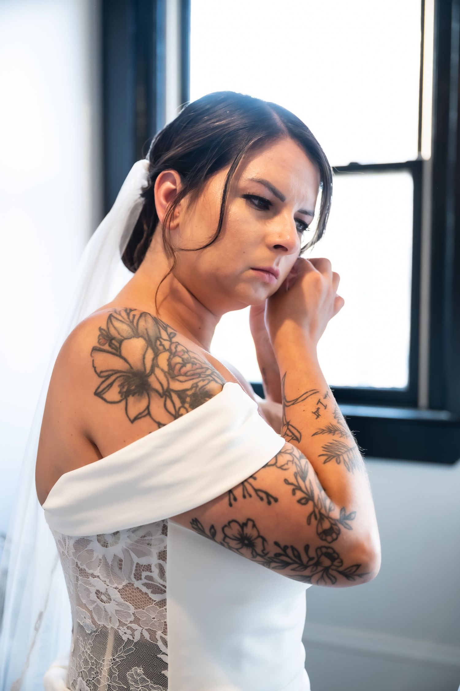 Tattooed bride in off-shoulder lace wedding dress and veil adjusting earring near window.