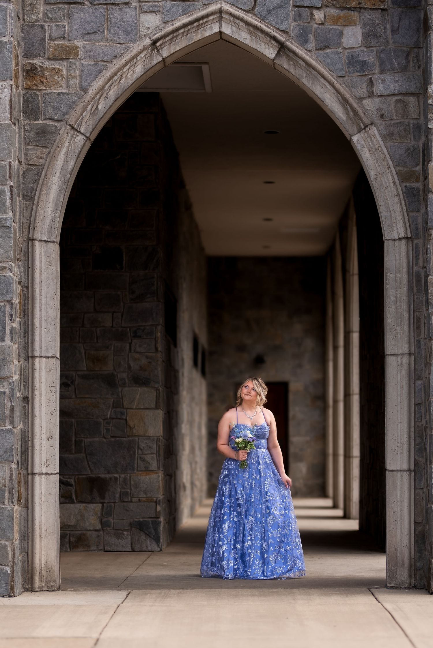 Woman in blue floral gown standing under gothic stone arch in castle corridor holding bouquet.