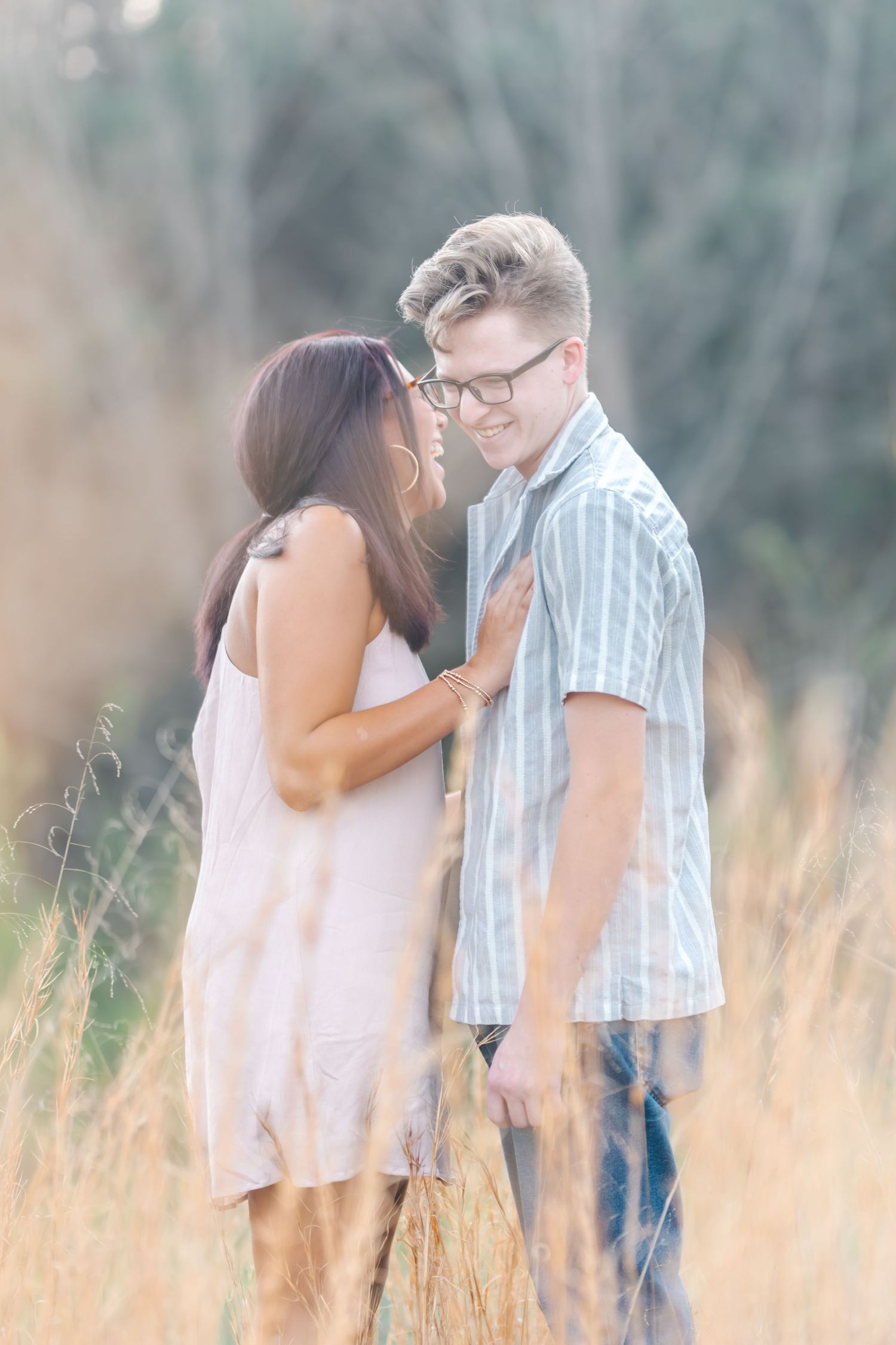 Couple smiling at each other in golden sunlit tall grass field during engagement session.