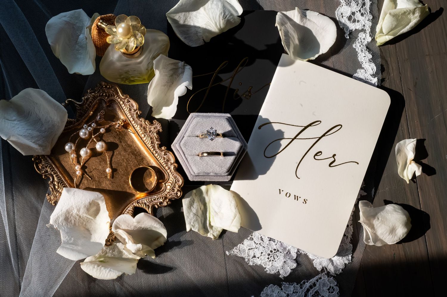 Elegant wedding flat lay with vow book, diamond rings, gold jewelry, pearl earrings, and white rose petals.