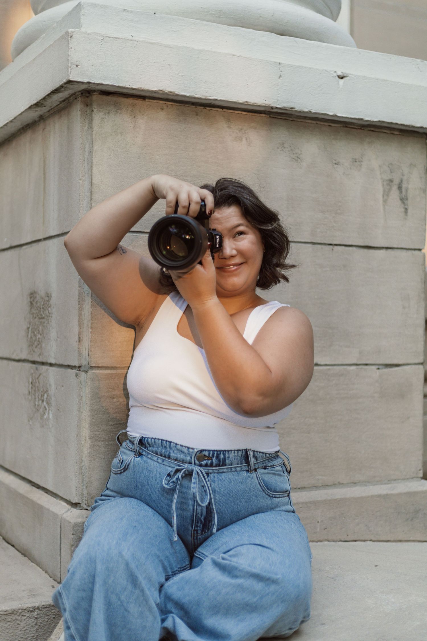 A plus-size woman in jeans and white tank top sits outdoors holding a DSLR camera up to photograph.