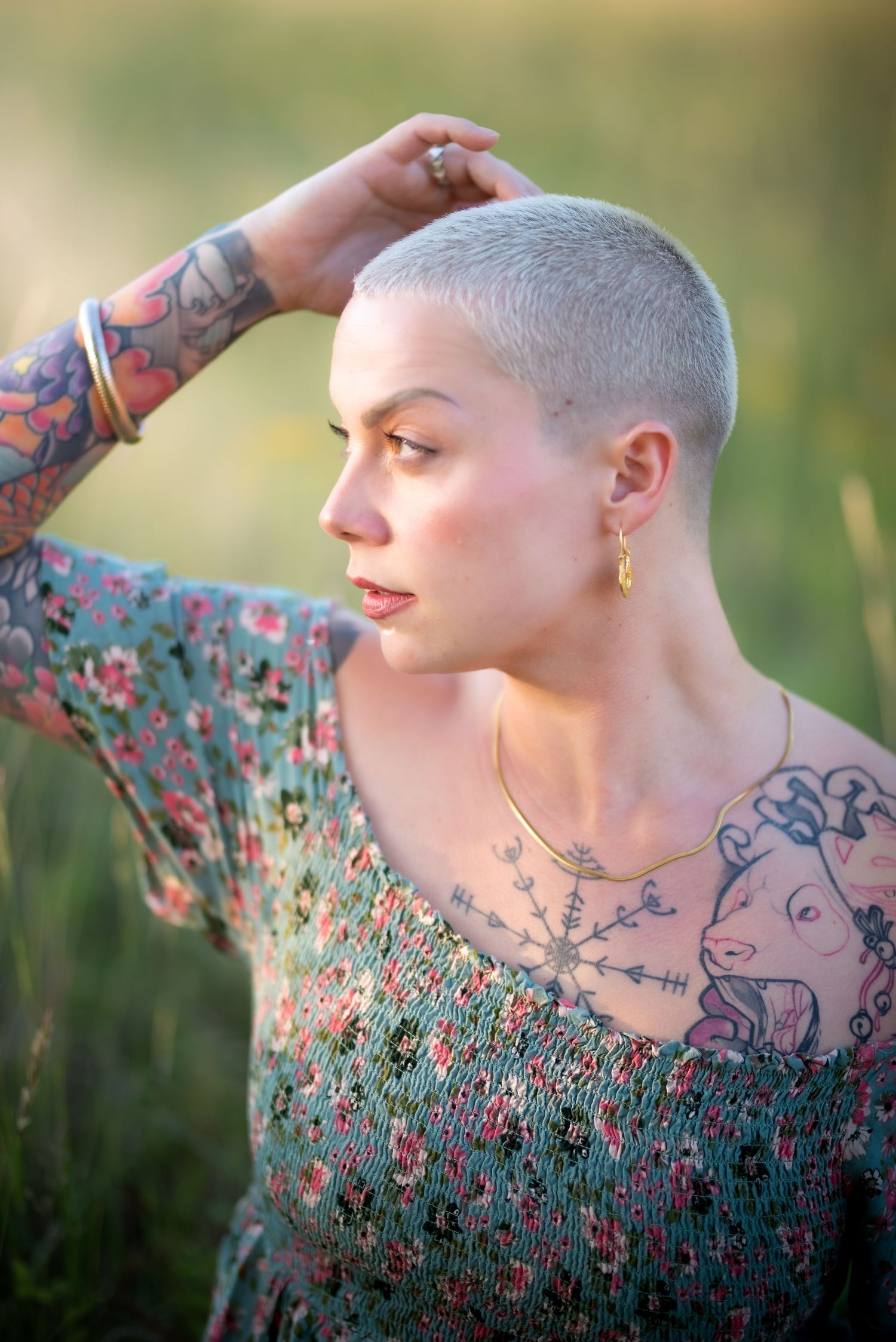 Woman with shaved head and tattoos posing outdoors in floral dress during golden hour photography.