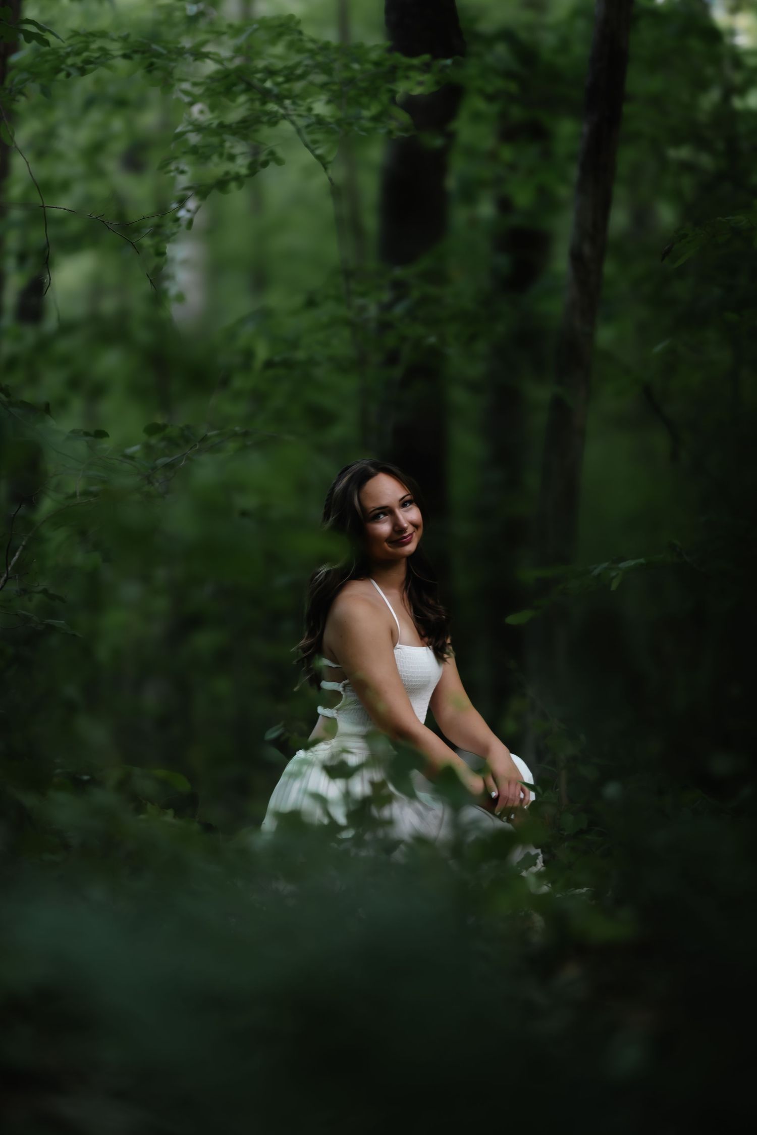 A smiling bride in a white dress stands among lush green forest trees, captured in a dreamy outdoor portrait.