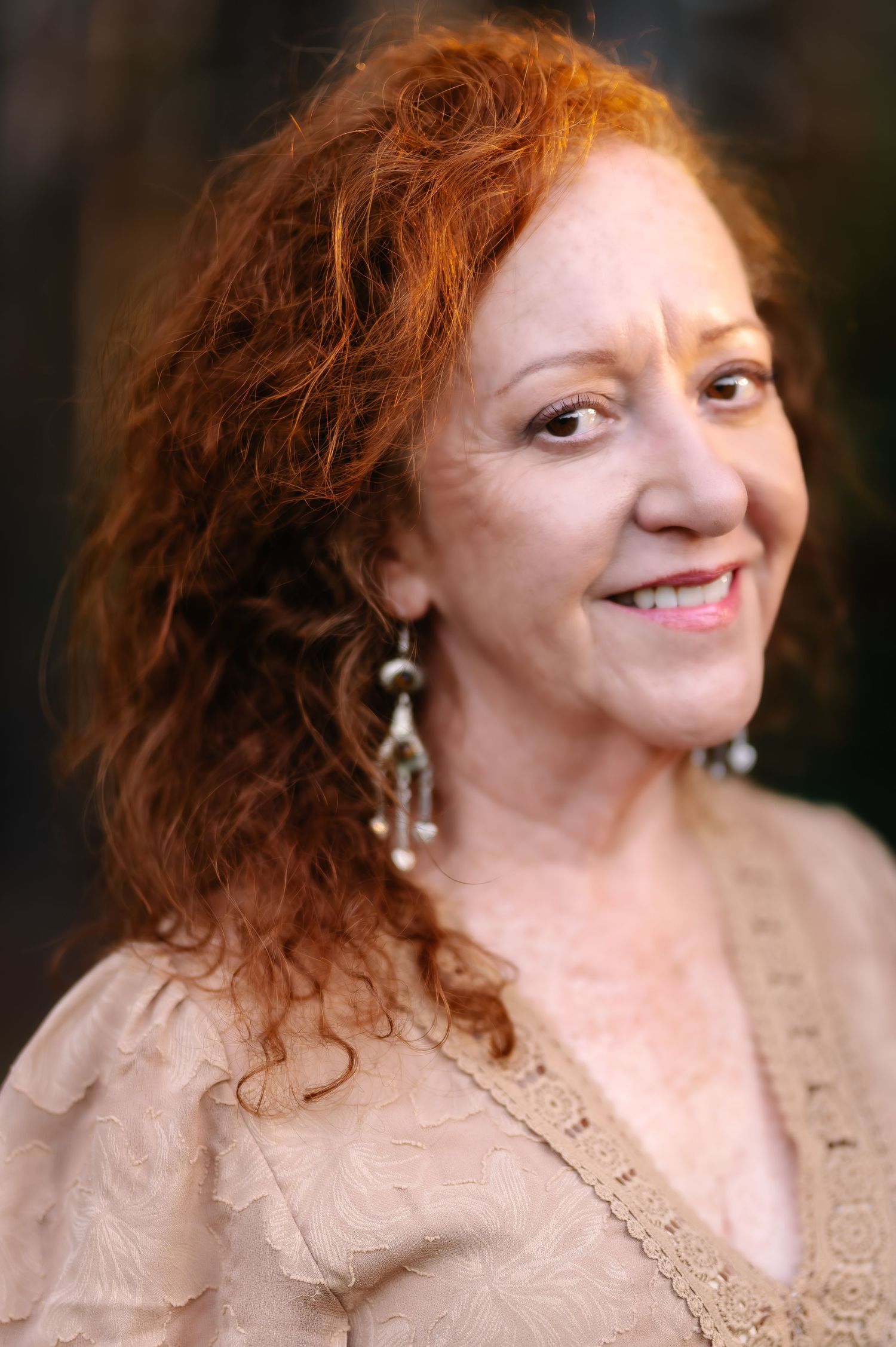 Smiling middle-aged woman with curly red hair wearing elegant drop earrings and a beige lace top.