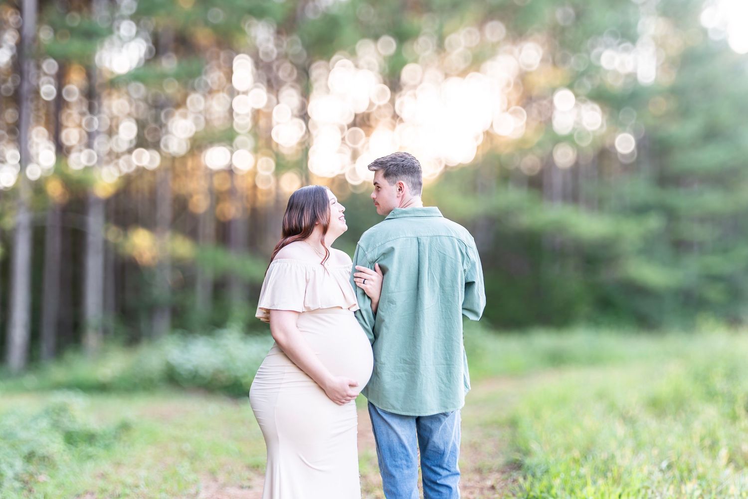 A pregnant couple walks hand in hand along a path near a sunlit pine forest.