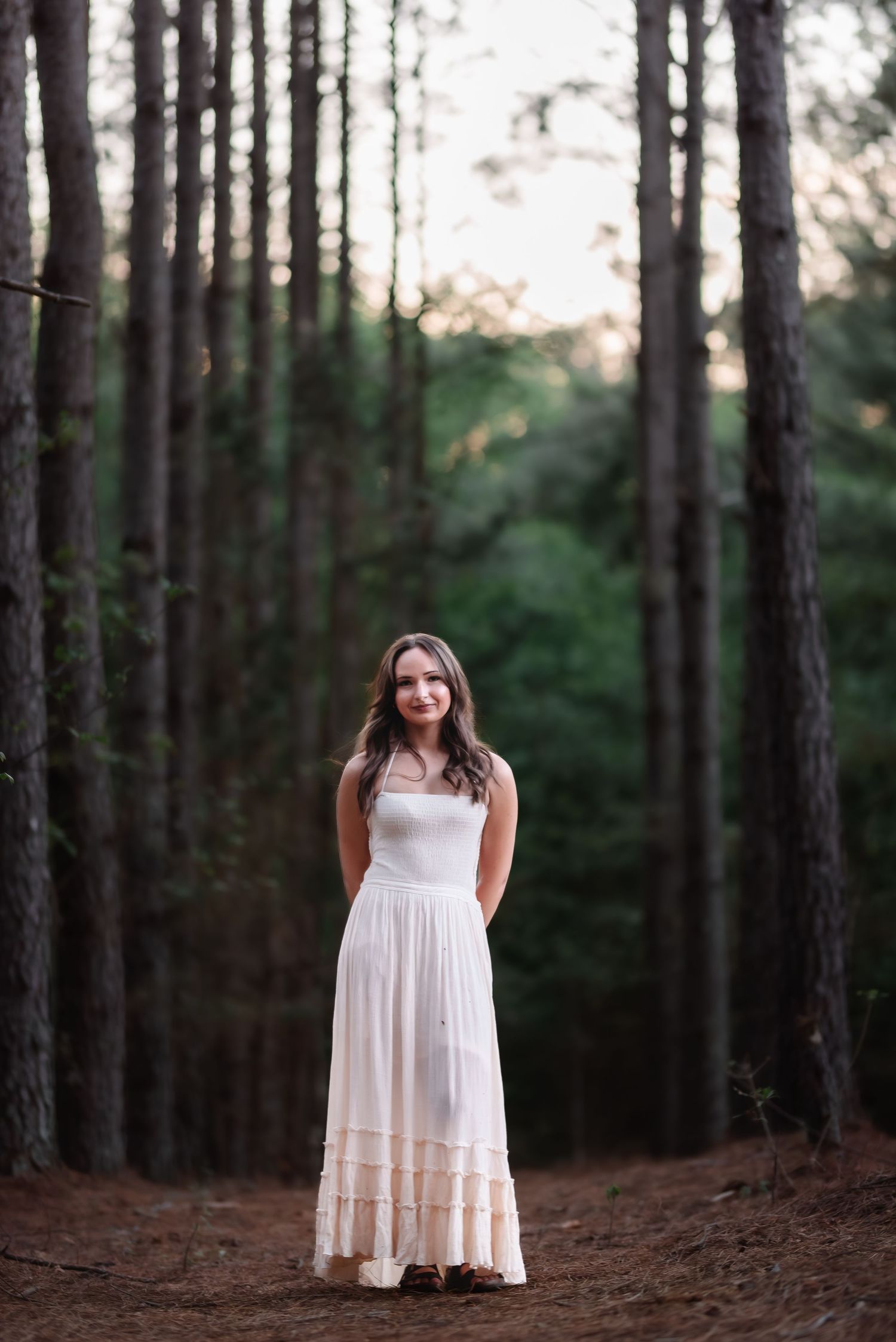 Young woman in white maxi dress standing among tall pine trees in a forest, smiling at camera.
