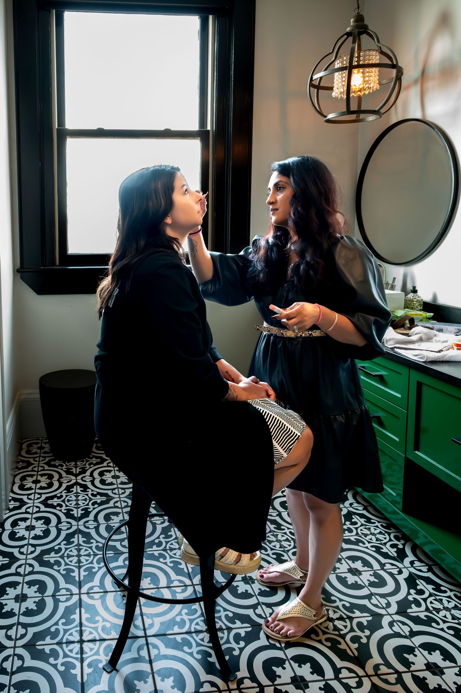 A makeup artist applies makeup to a seated woman in a stylish bathroom with patterned tile floors and green cabinetry.