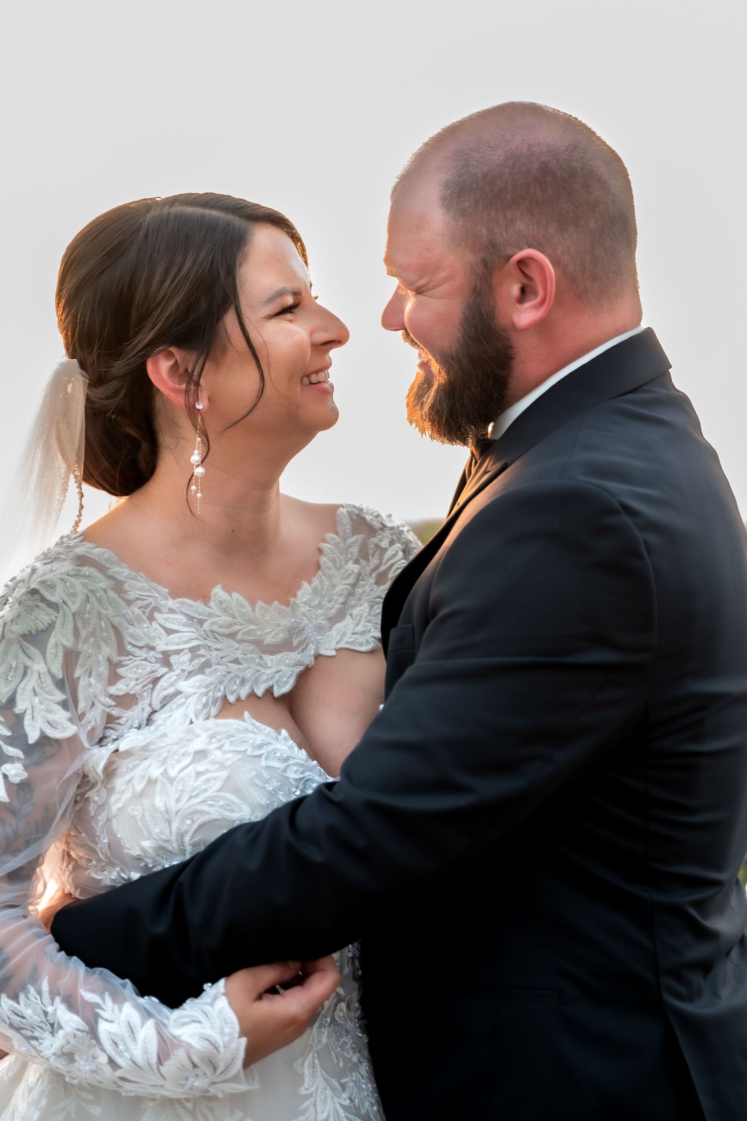 A smiling bride in lace gown and groom in black suit share a tender moment, gazing at each other on their wedding day.