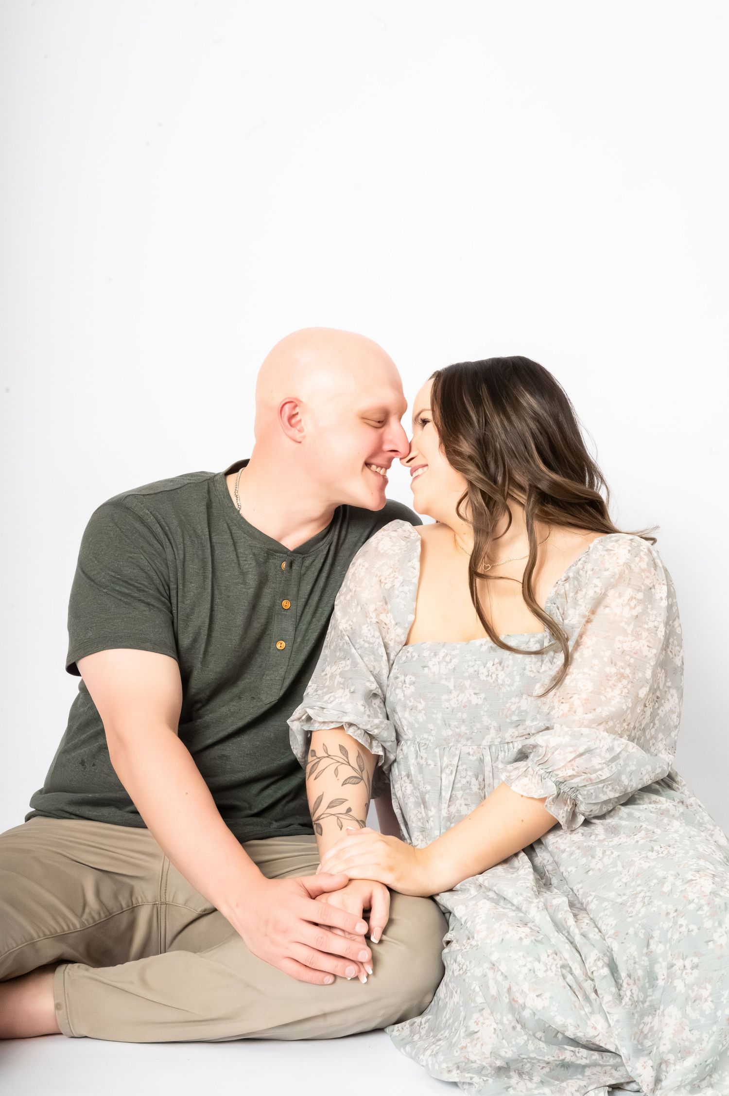 A bald man and brunette woman in floral dress sit on white floor, smiling and leaning in for a kiss.