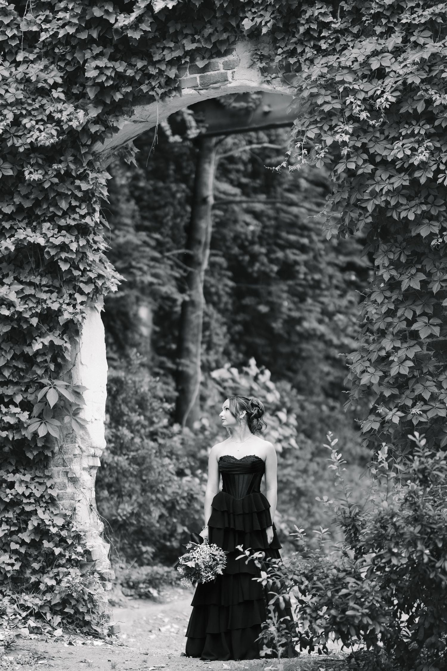 Black and white photo of a woman in a dark strapless gown holding flowers, standing beneath an ivy-covered stone archway.