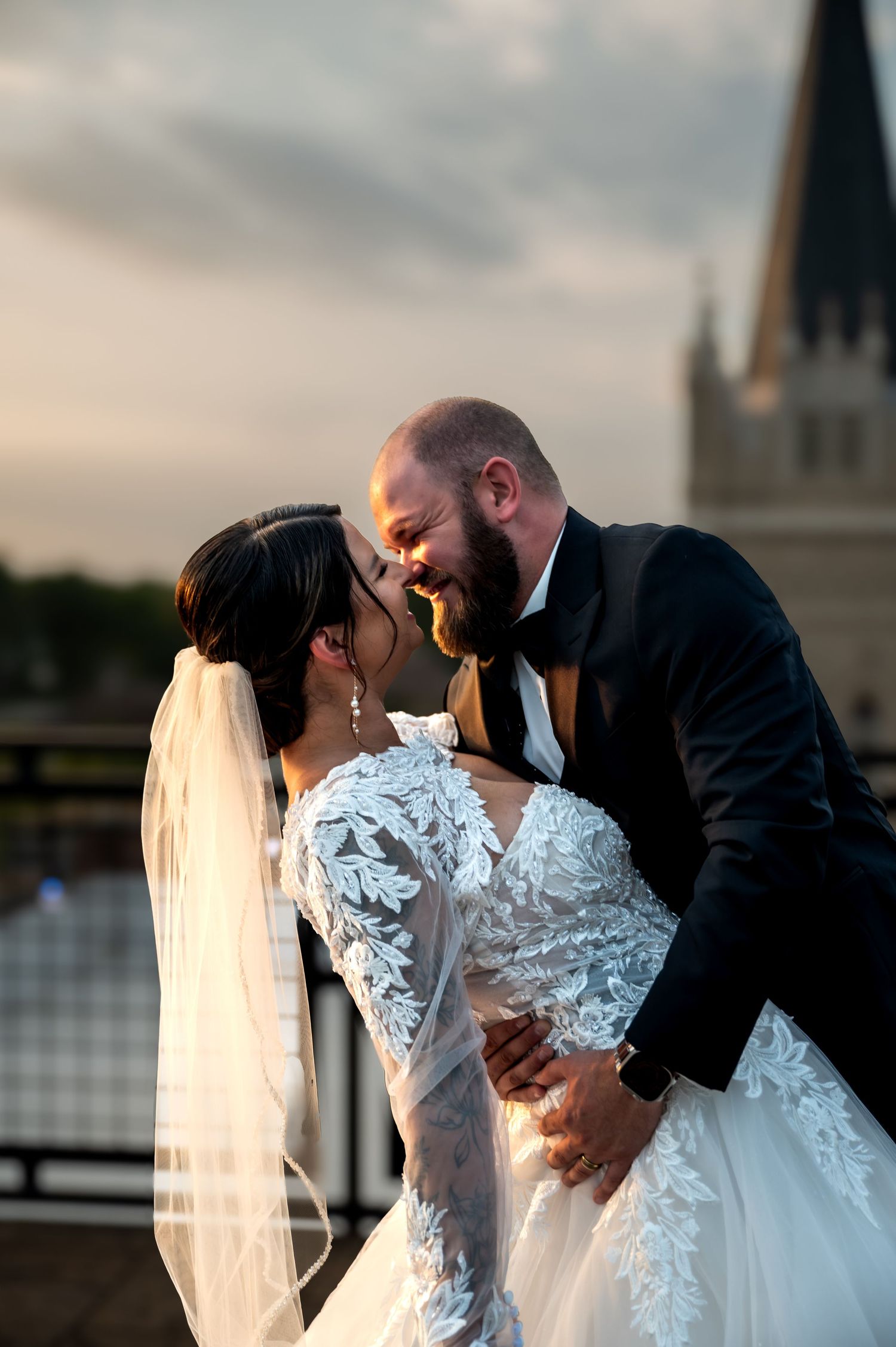 Bride and groom share a romantic moment, nearly kissing outdoors at golden hour, bride in lace gown with veil.